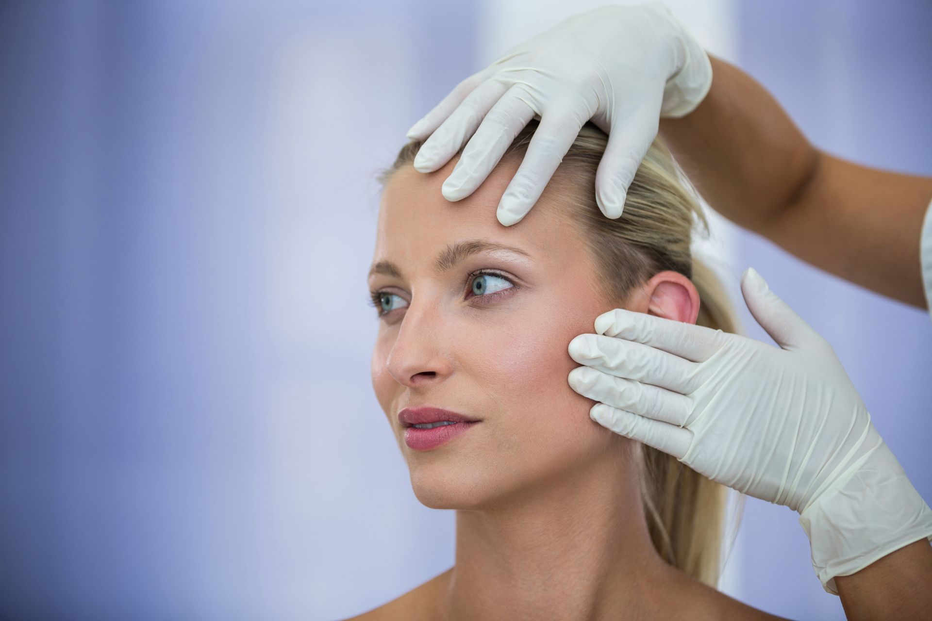 Woman's face examined by gloved hands, possibly a medical checkup, in a clinic setting.