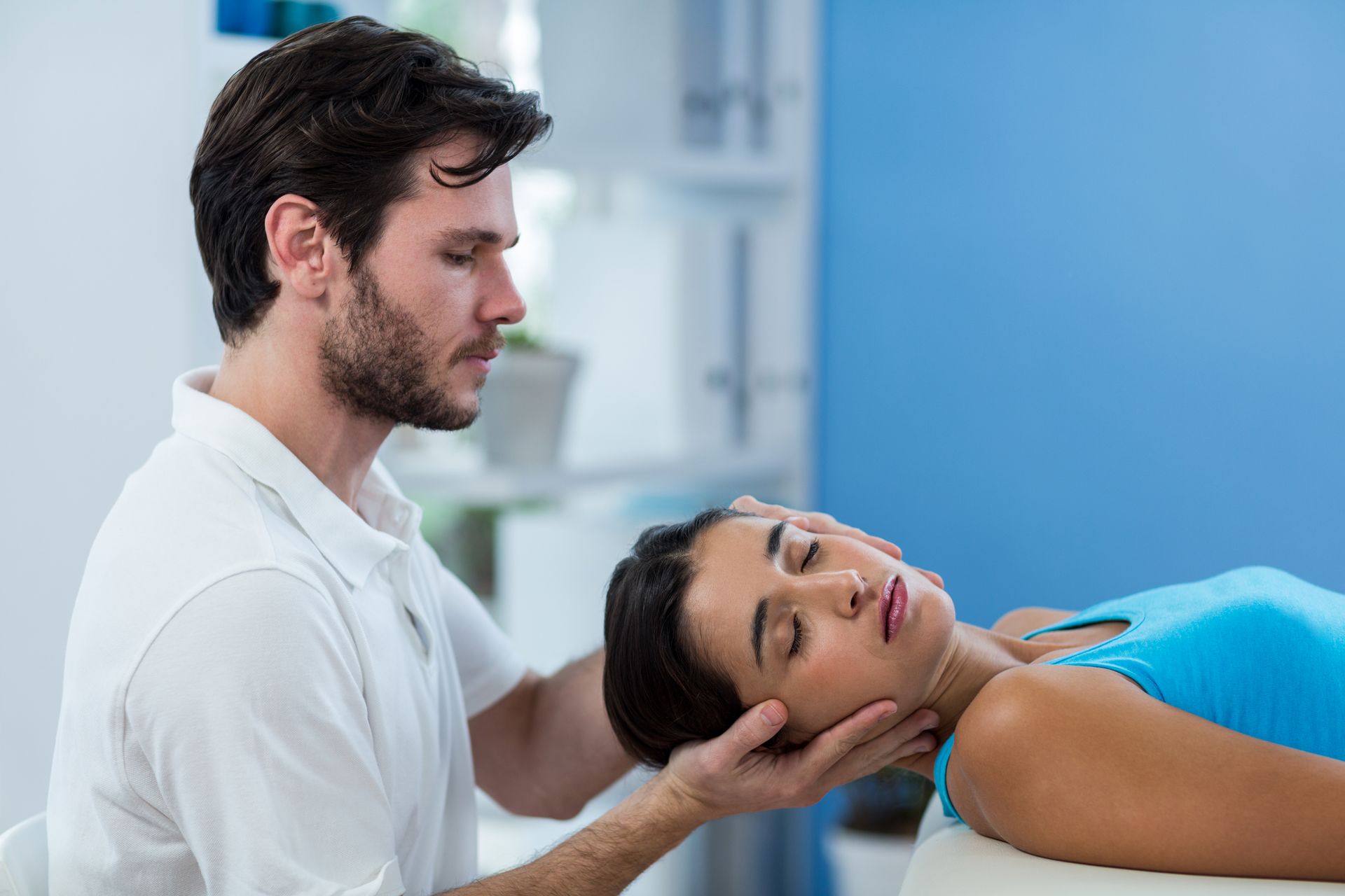 Man giving neck therapy to a woman lying down. Blue wall.