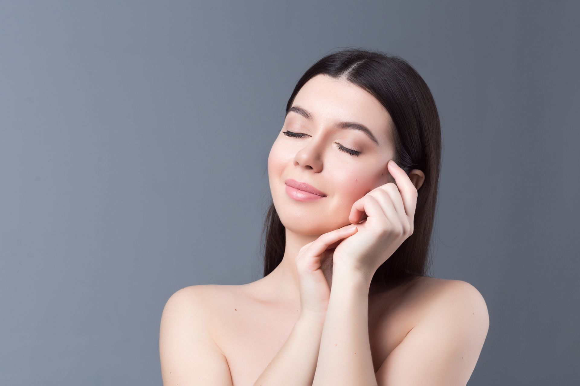 Woman with eyes closed, gently touching her face, smiling; gray background.