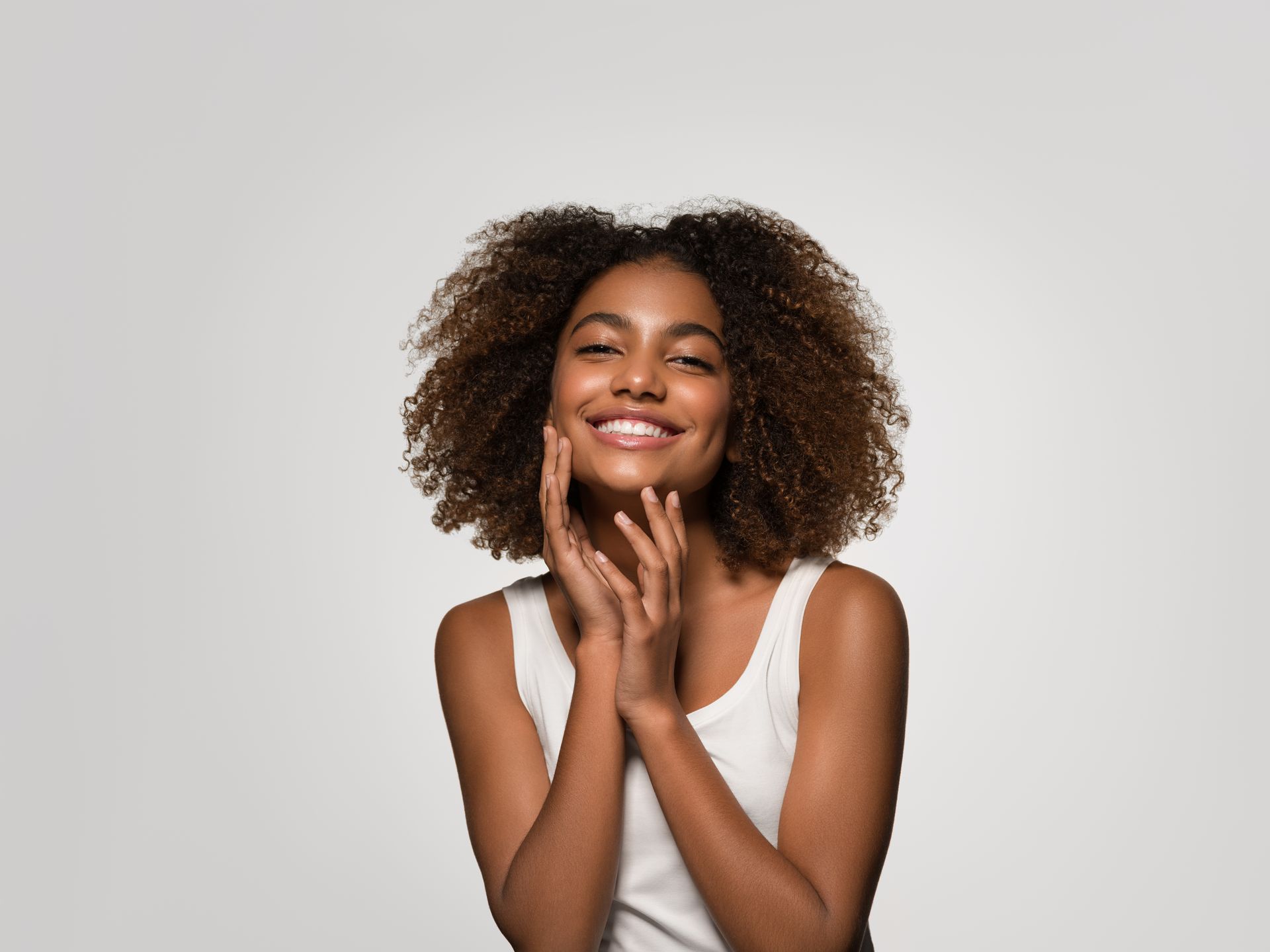 A woman with curly hair is smiling and touching her face.