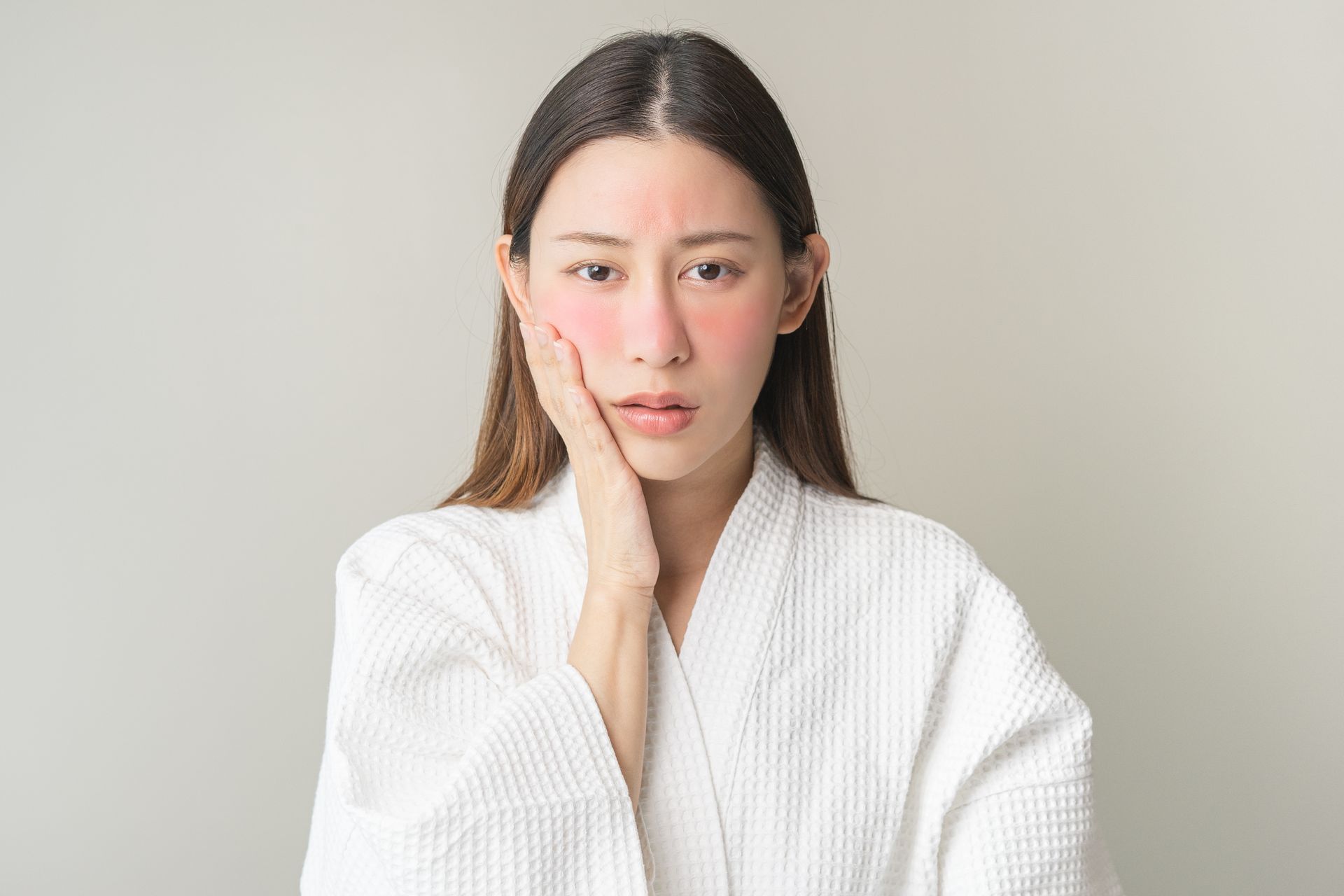 Woman in white robe, with red, flushed cheeks, touching her jaw, looking concerned.