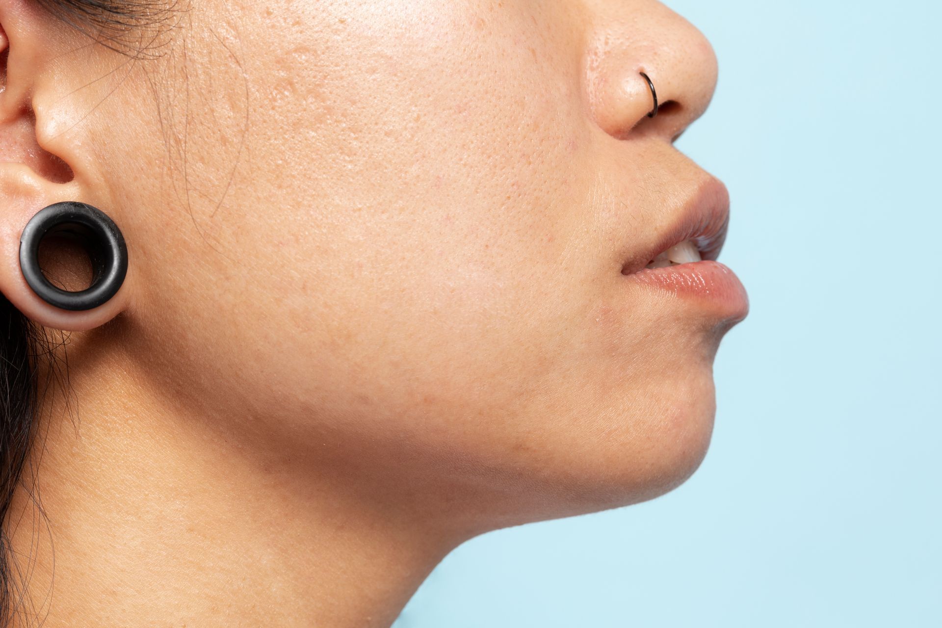Person's profile: pierced nose, earlobe gauge, light skin, looking up with slightly parted lips, on blue backdrop.