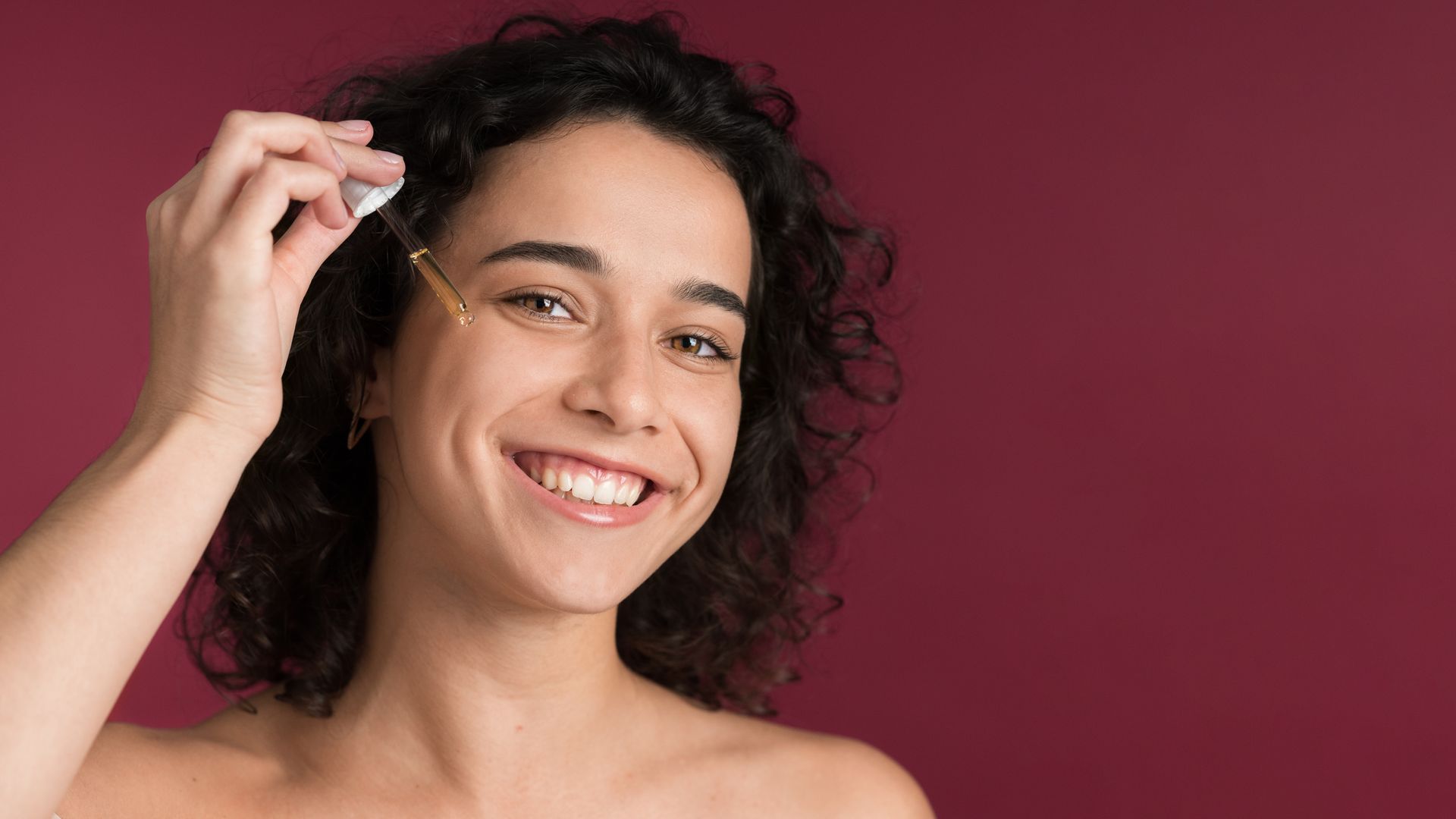 A woman is smiling while applying a serum to her face.
