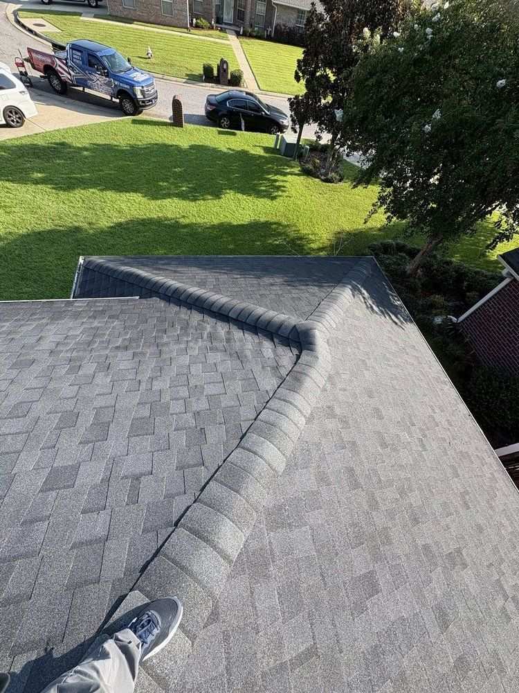 Rooftop with grey shingles, a person's foot, green lawn, and vehicles in the background.