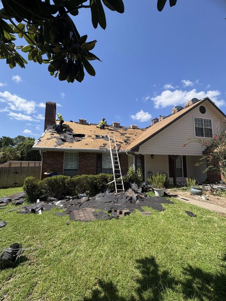 Roofers replacing a roof on a two-story house, debris on lawn, ladder, clear blue sky.