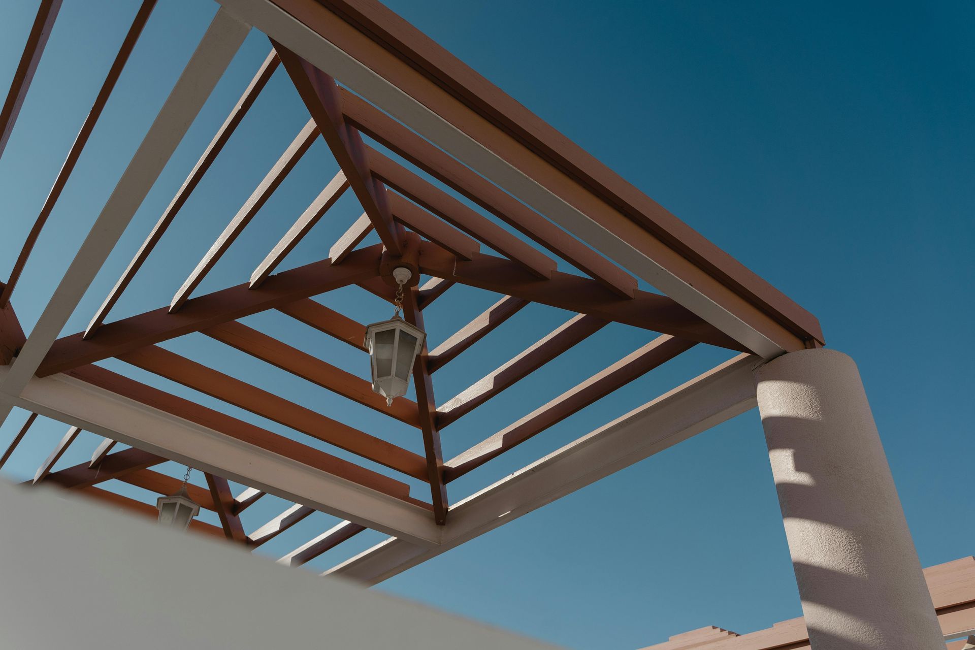 Wooden pergola with a hanging light fixture, set against a clear, blue sky. White pillars and walls are visible.