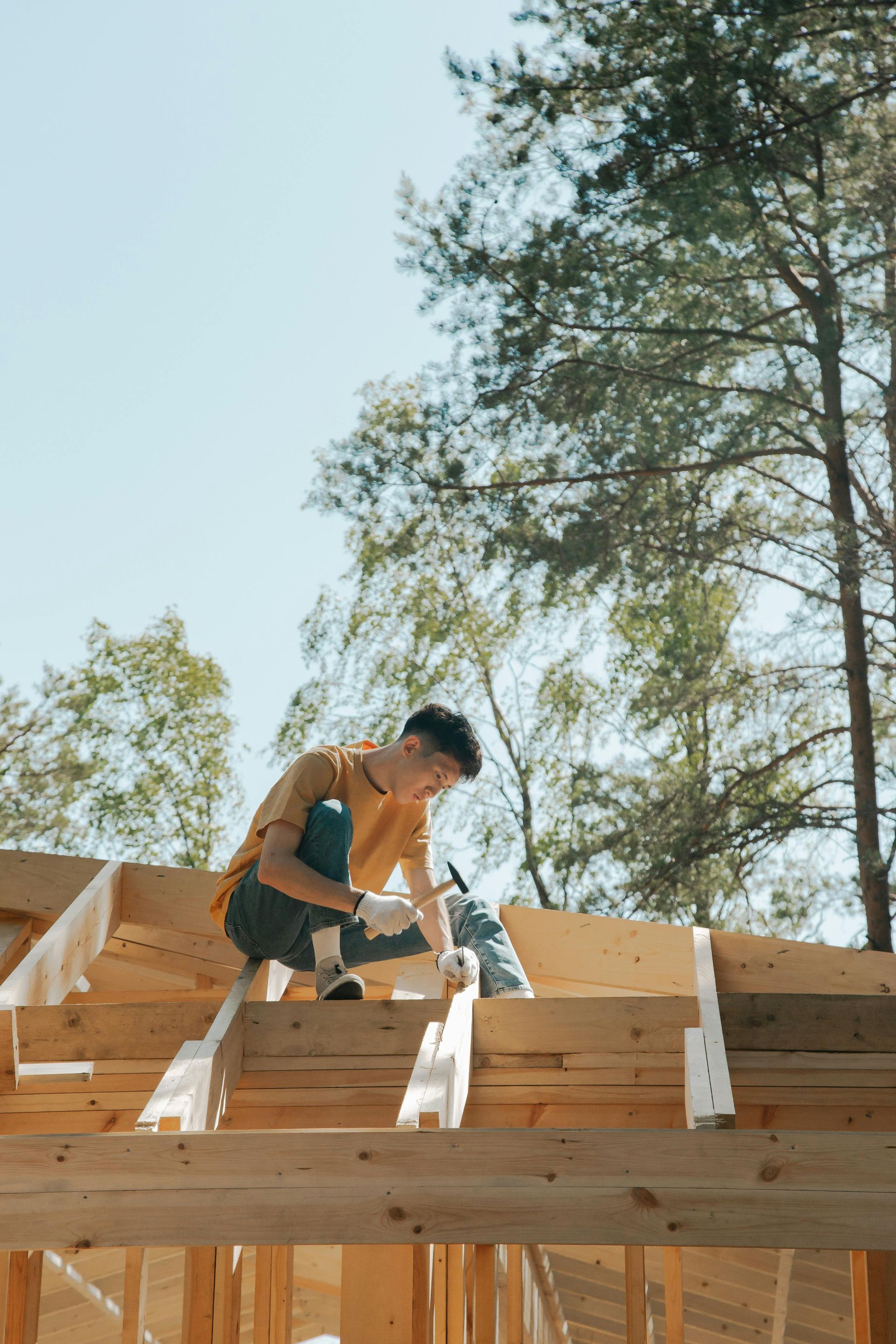 A person in a yellow shirt, wearing gloves, building on a wooden roof structure outdoors, surrounded by trees.