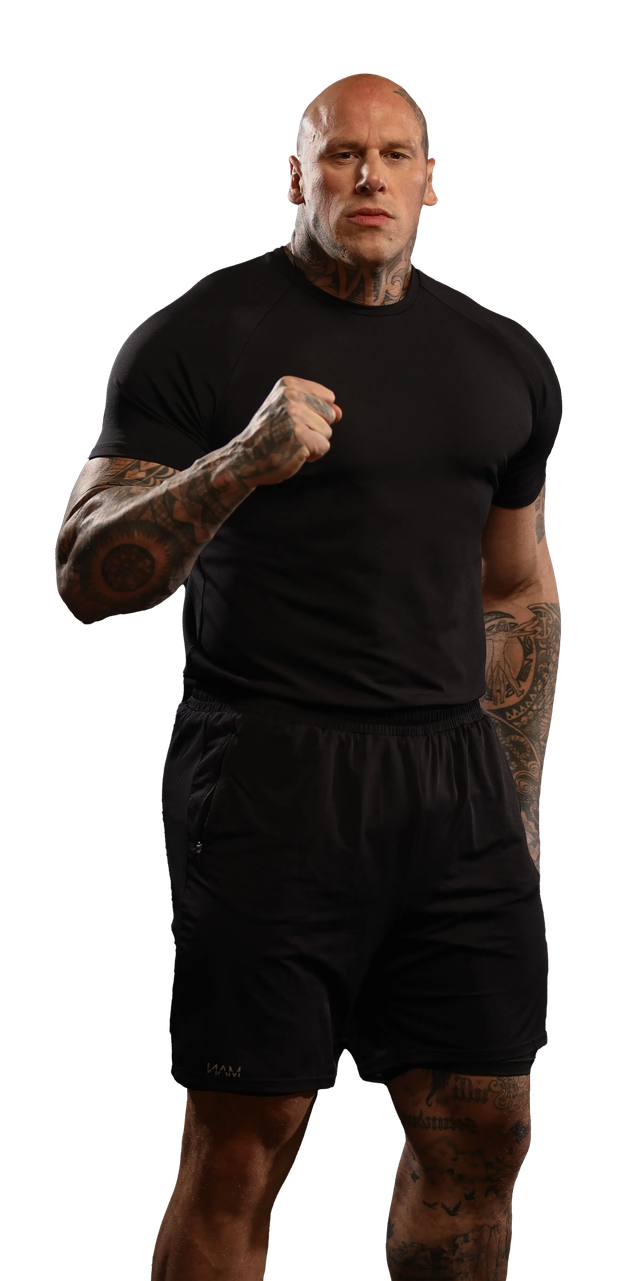 A muscular man in a black shirt and shorts, fist clenched, looks forward.