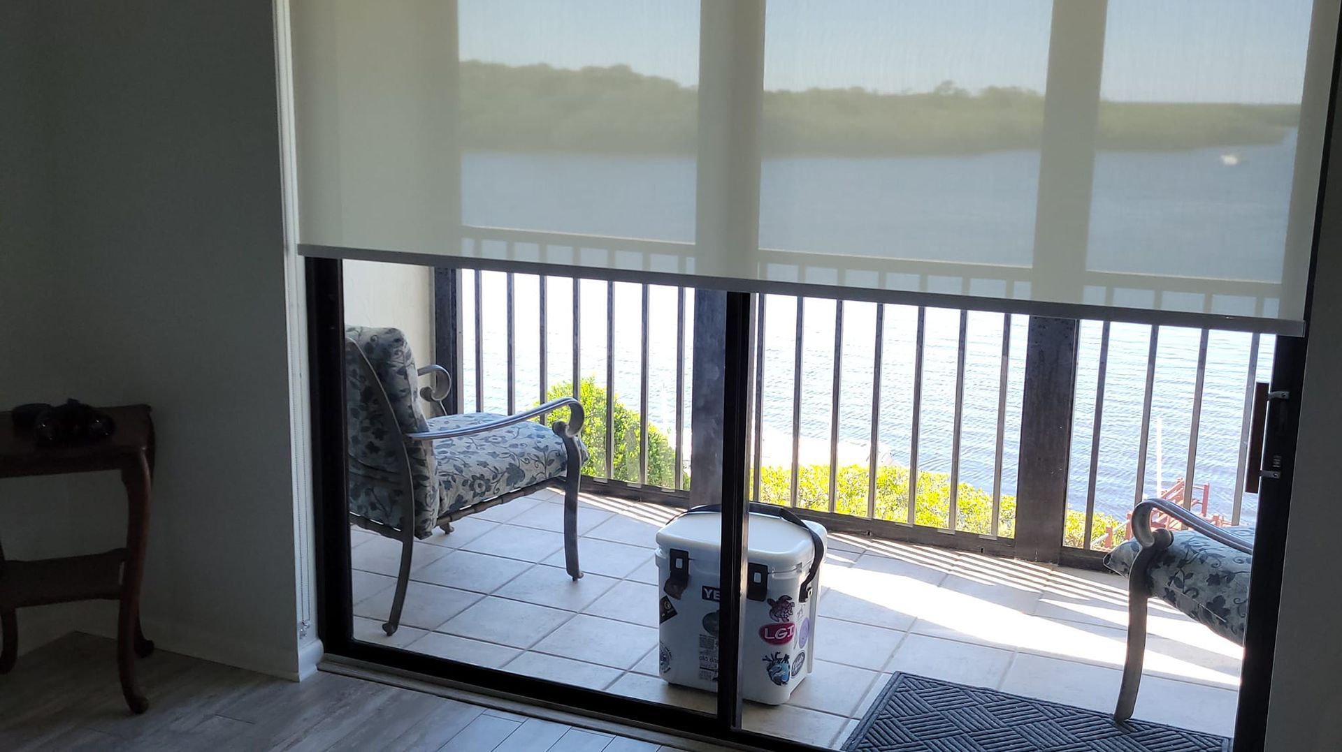 Balcony overlooking water, with chairs, trash can, and blinds partially covering windows.