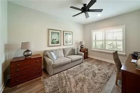 Cozy home office with sofa, desk, rug, and art. Neutral tones, natural light, ceiling fan.