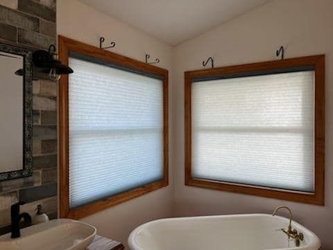 Bathroom corner with two windows, each with beige cellular shades and wooden frames, above a white bathtub.