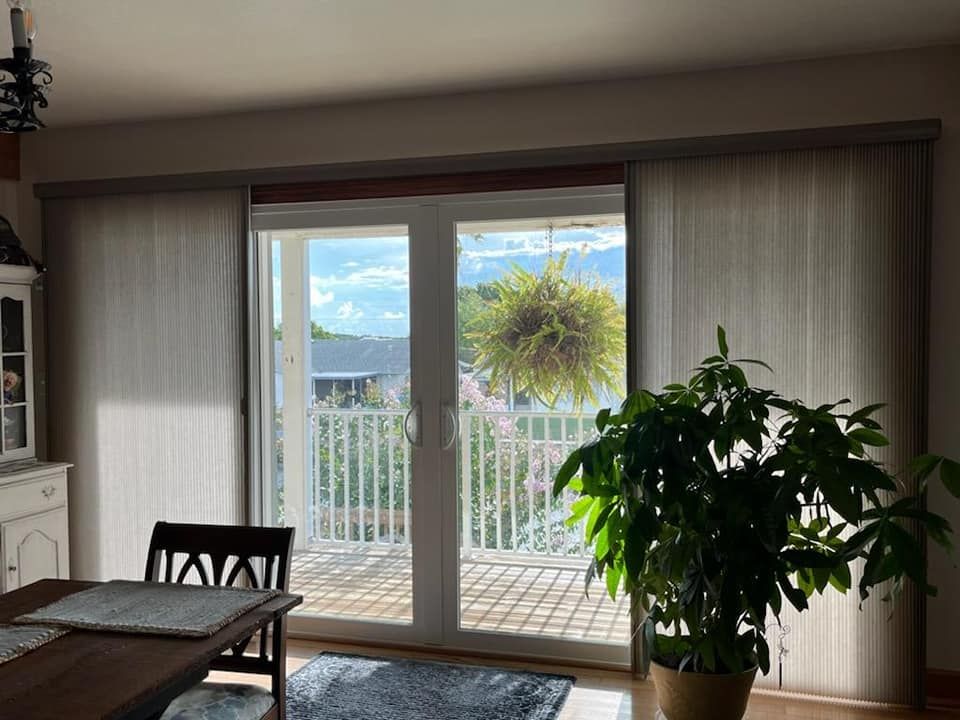 Interior view of a room with sliding glass doors, sheer blinds, and a large plant. Outside, a balcony and mountain views.