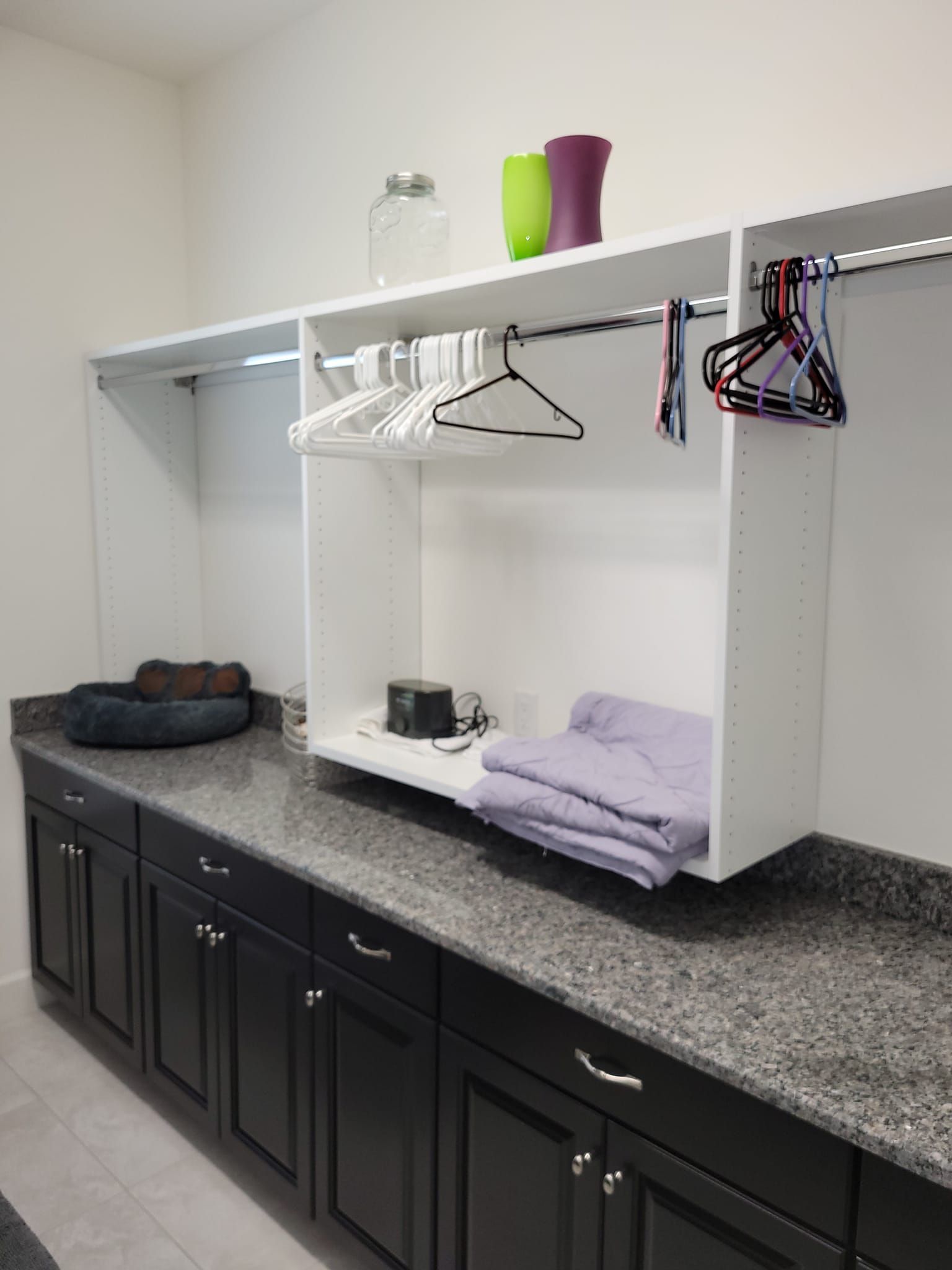 Laundry room with dark cabinets, gray countertop, and white shelving with clothes hanging.