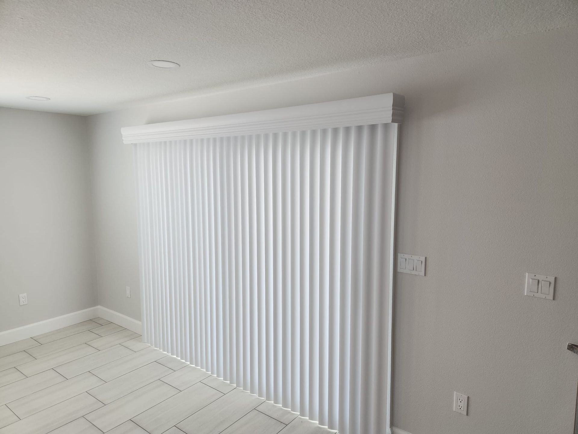 White vertical blinds covering a large sliding glass door, in a room with light grey walls and tile flooring.