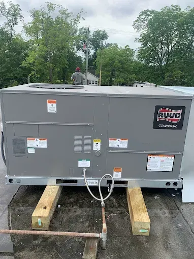 Rooftop HVAC unit on wooden supports, with Ruud logo. Trees in the background. Gray and silver.