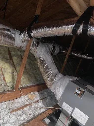 HVAC unit and ductwork in an attic, with insulation and wooden beams visible.