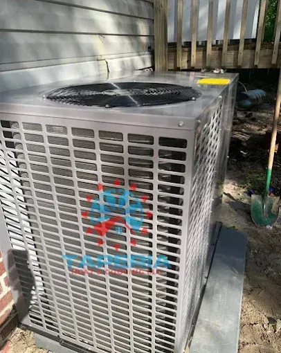 New gray air conditioning unit on a concrete pad next to a house with a shovel in the background.