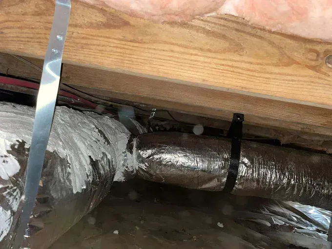 Metallic ductwork secured with black zip tie and metal strapping under wooden beam with insulation.