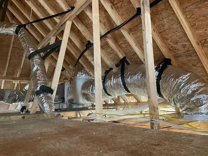 HVAC ductwork in an attic, supported by wooden beams. Silver insulated tubes are held by black straps.