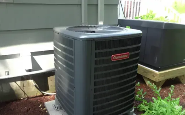 Goodman air conditioner unit outside a building, surrounded by mulch and greenery.