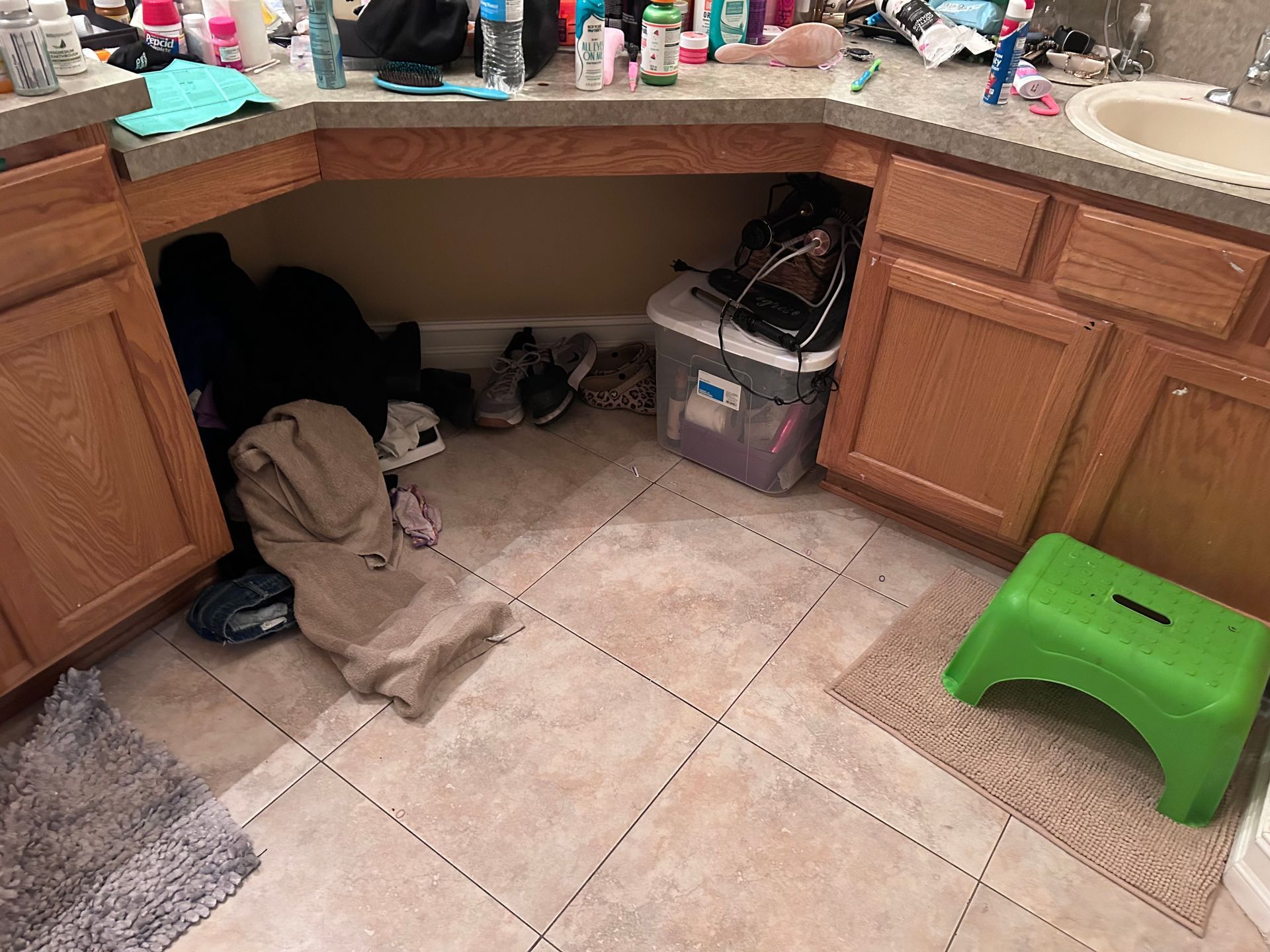 Bathroom with clutter: clothes, shoes, a clear bin, and a green step stool under the sink.