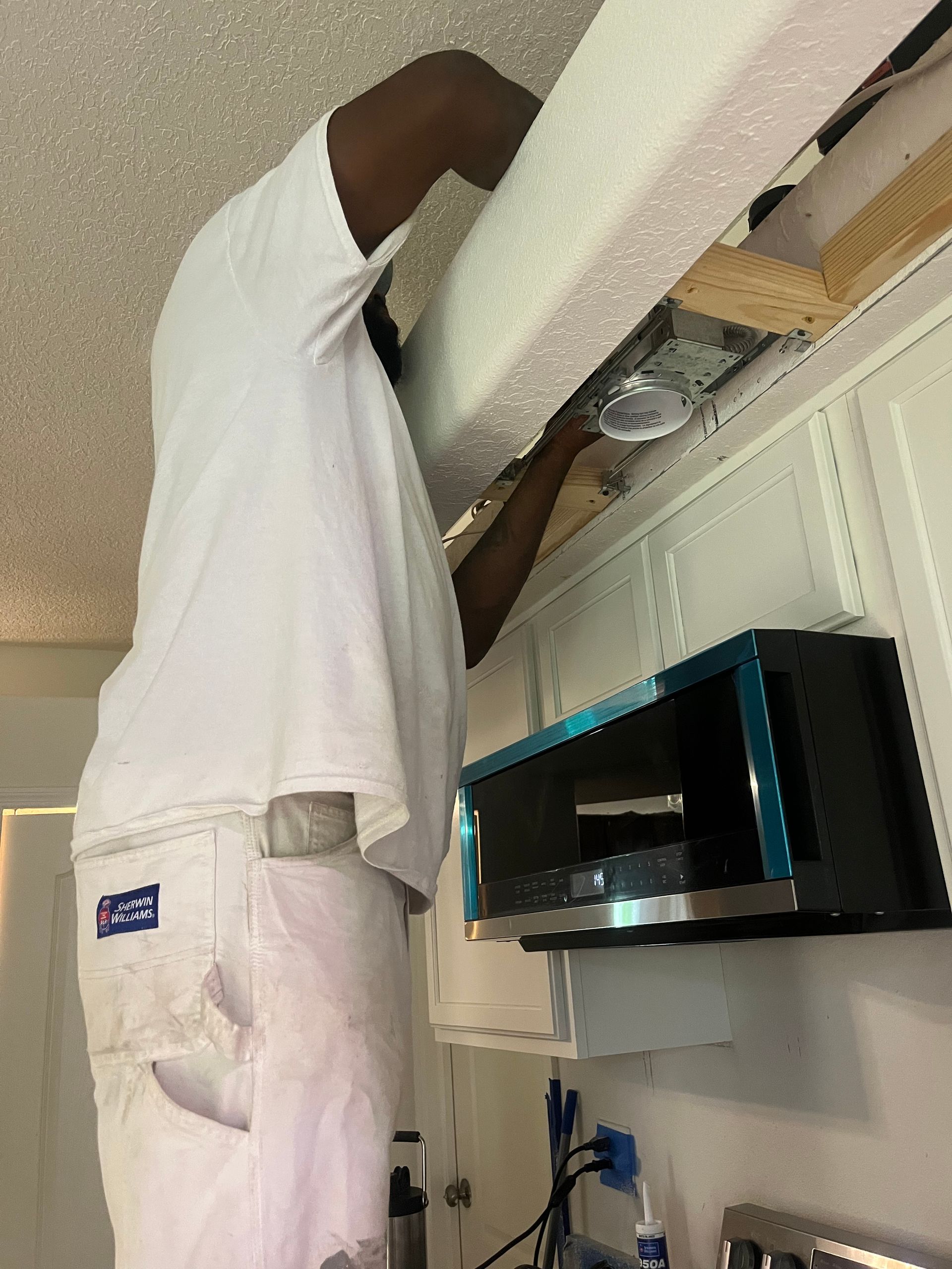 A man in white pants is working on a microwave in a kitchen.