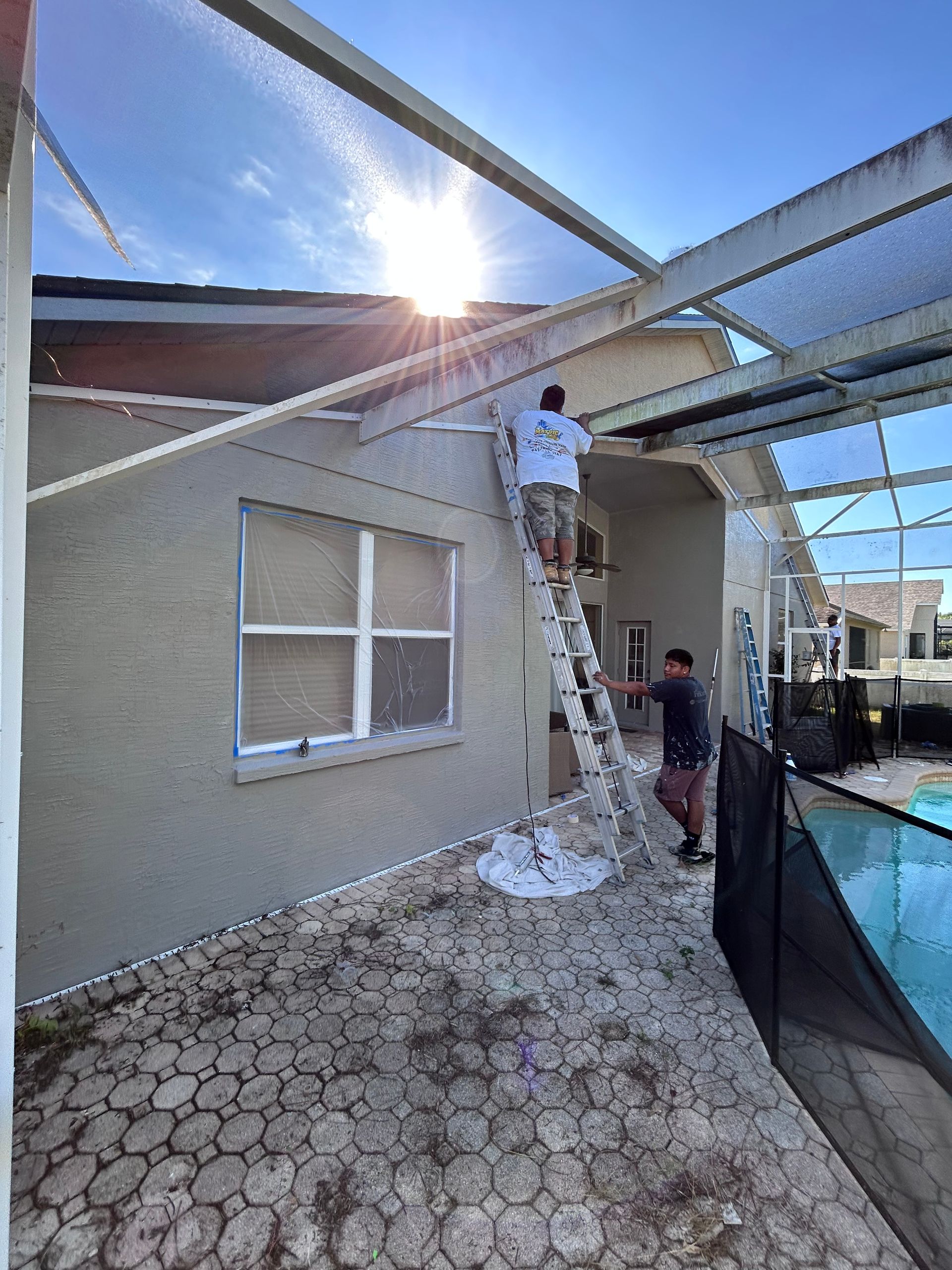 Two men on a ladder repair a white screen enclosure attached to a house with a pool, bright sun.