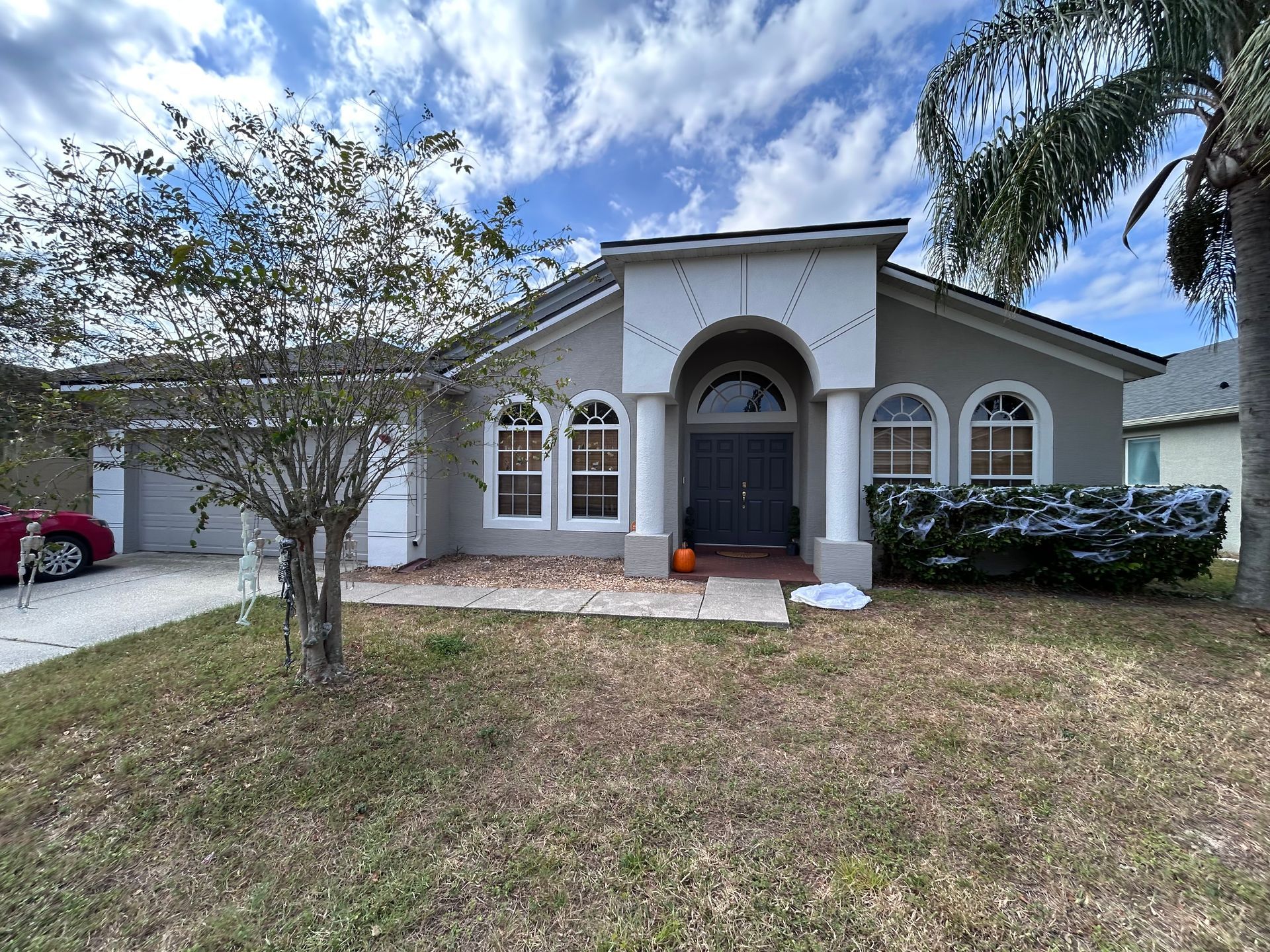 Suburban house with gray exterior, arched entry, two-car garage, and dry grass lawn on a sunny day.