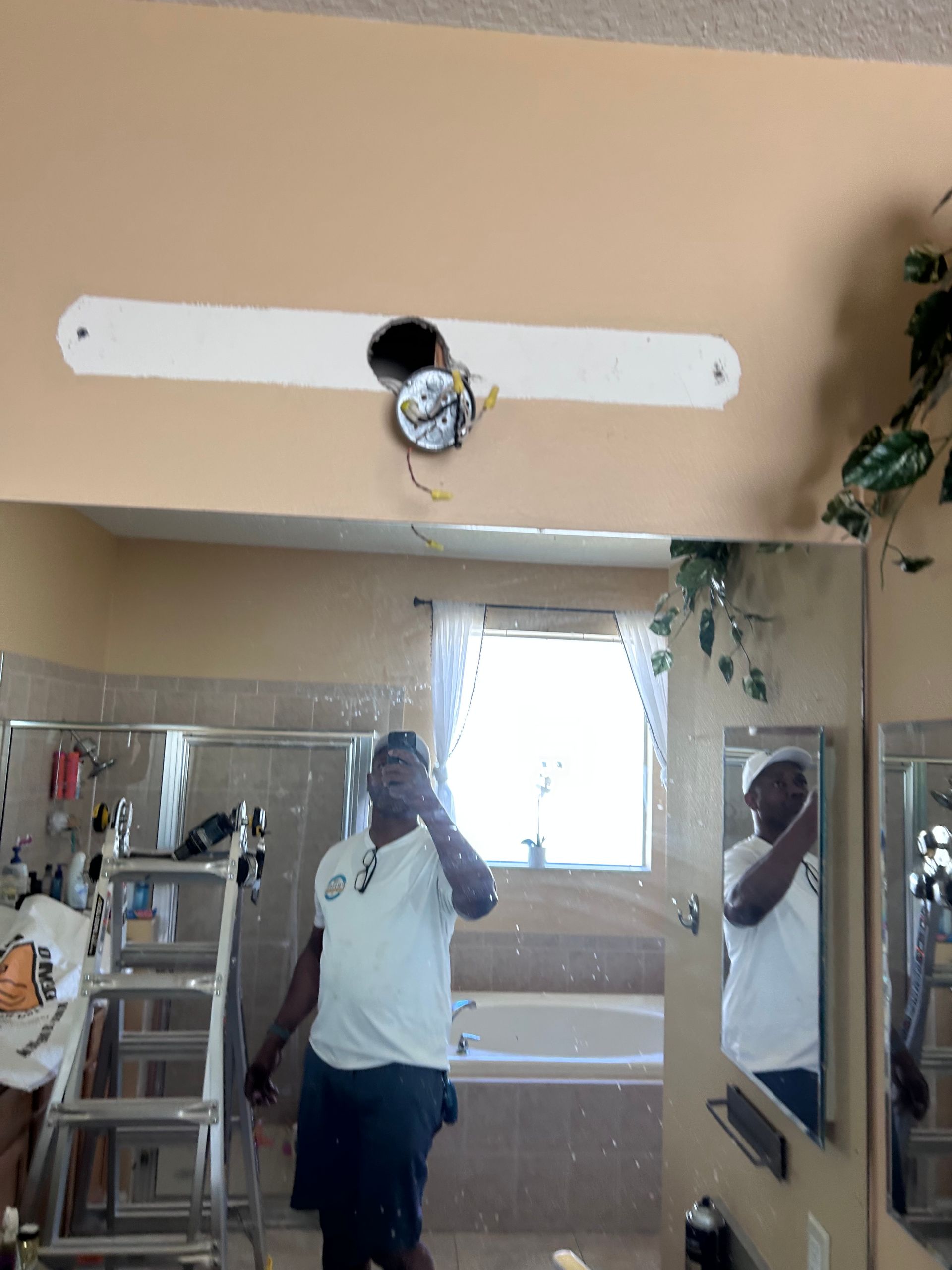 Person installing a light fixture above a bathroom mirror. A ladder and bathtub are visible.