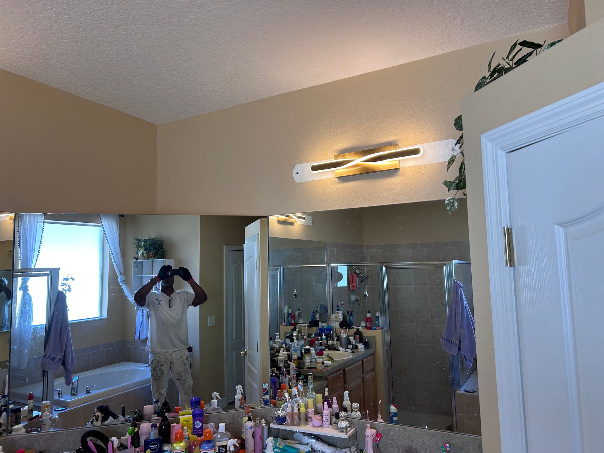 A bathroom with a large mirror reflecting a person and toiletries. A light fixture is mounted above the mirror.