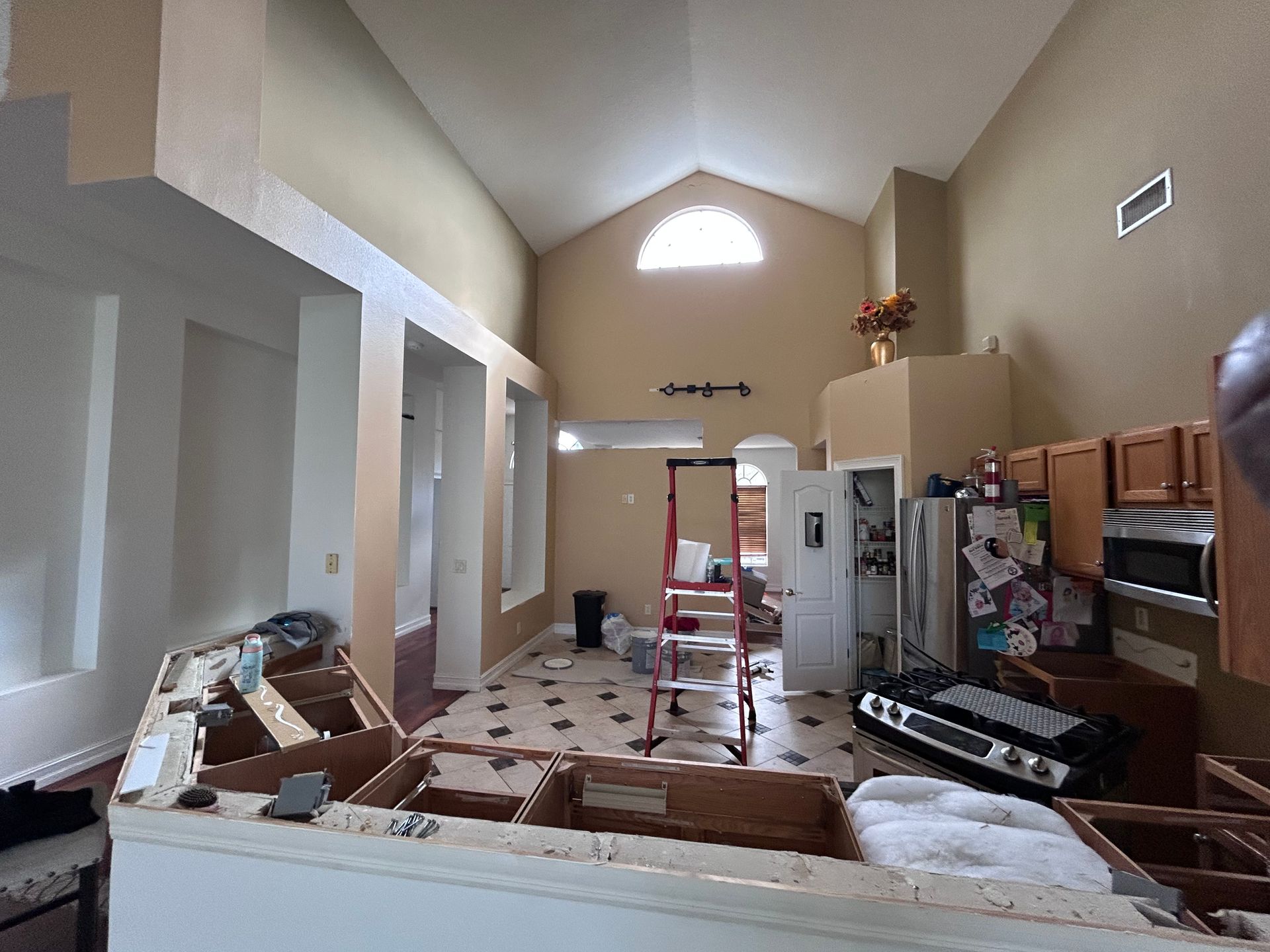 Kitchen undergoing renovation: cabinets removed, walls beige, high ceiling with arched window.