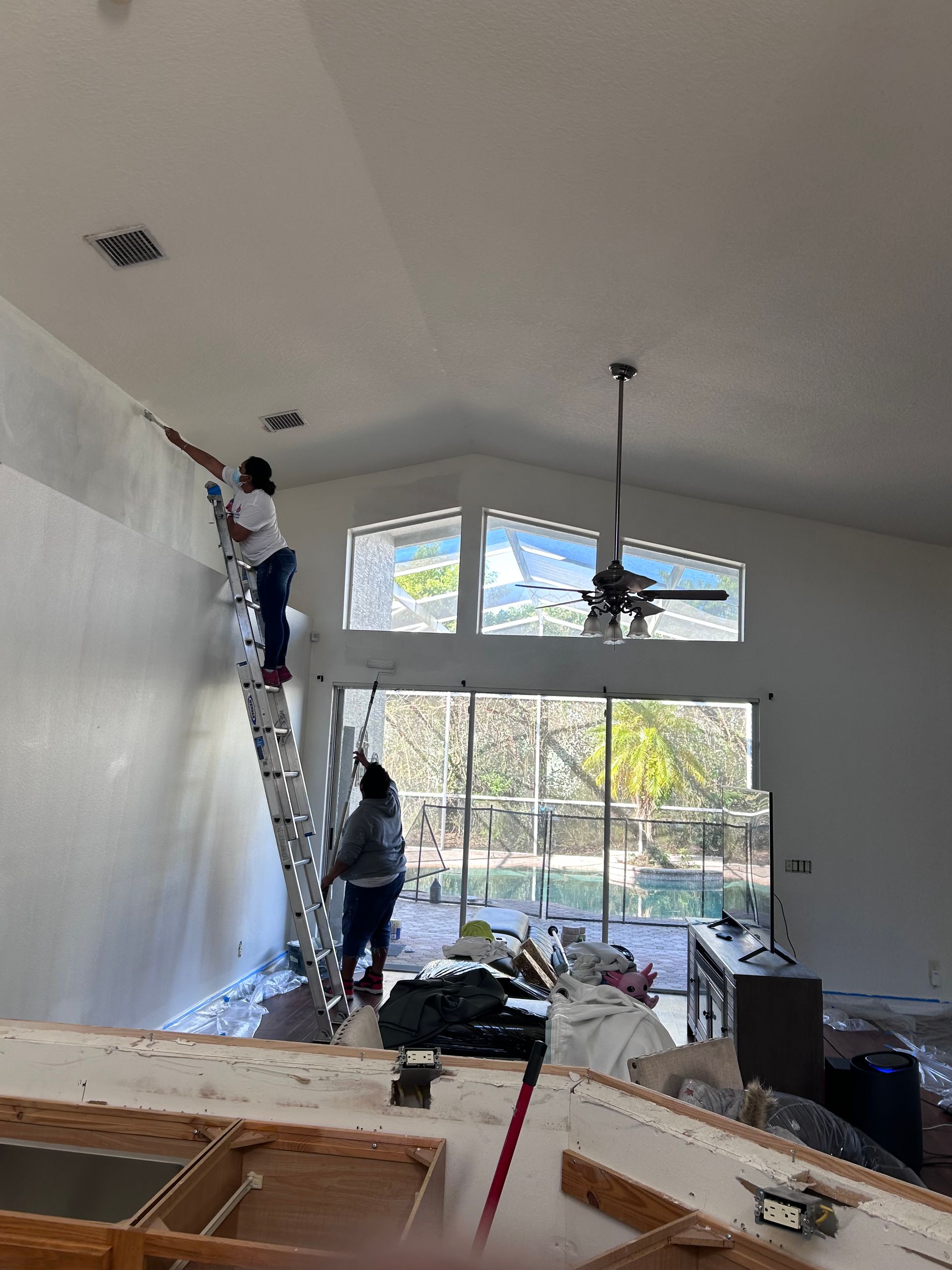Two people on ladders painting a tall white wall indoors.