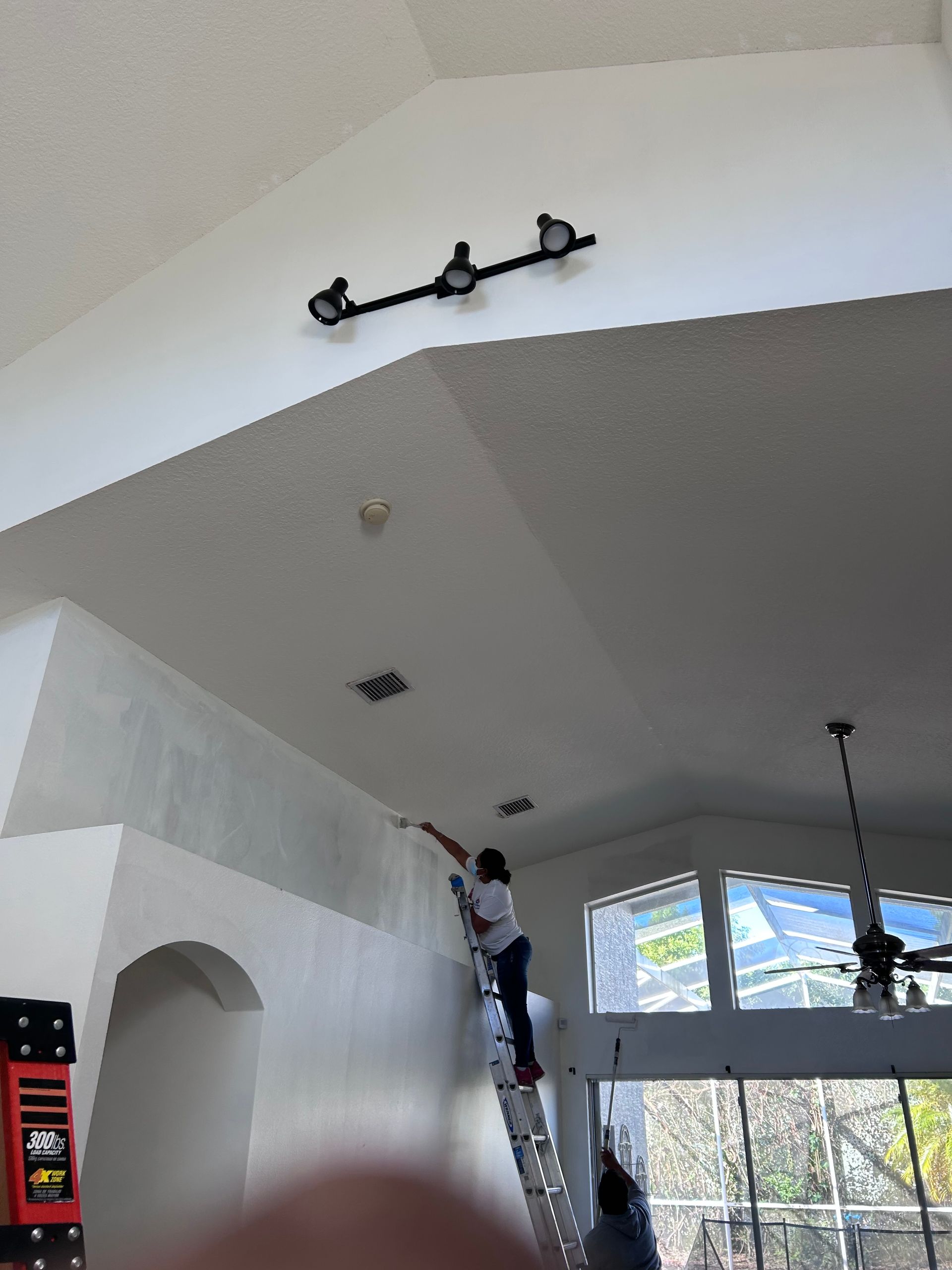 Person painting a high ceiling from a ladder indoors; light fixtures, arched doorway, and windows present.