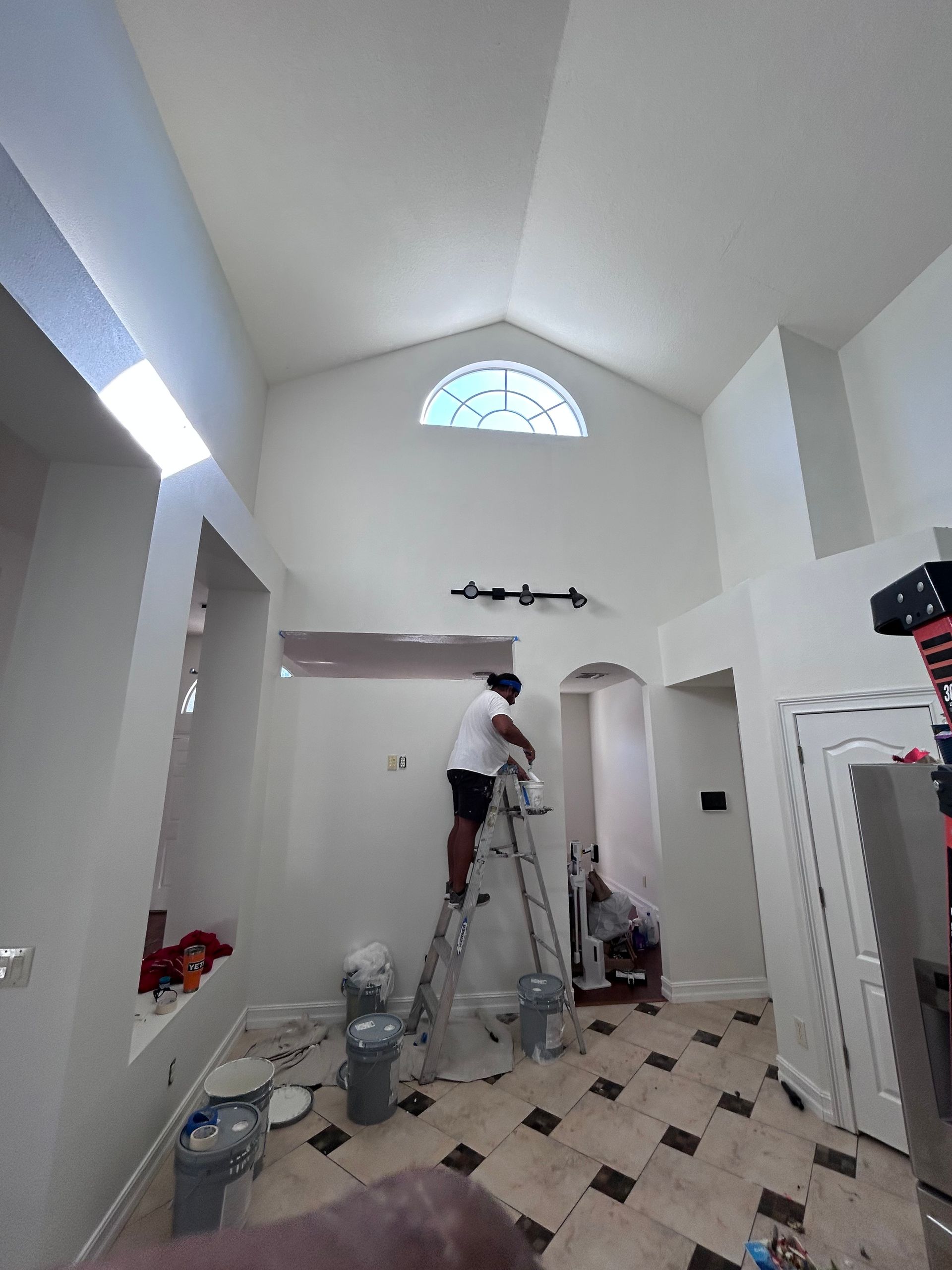 Person on a ladder painting a high white wall in a room with tiled floors and a window.