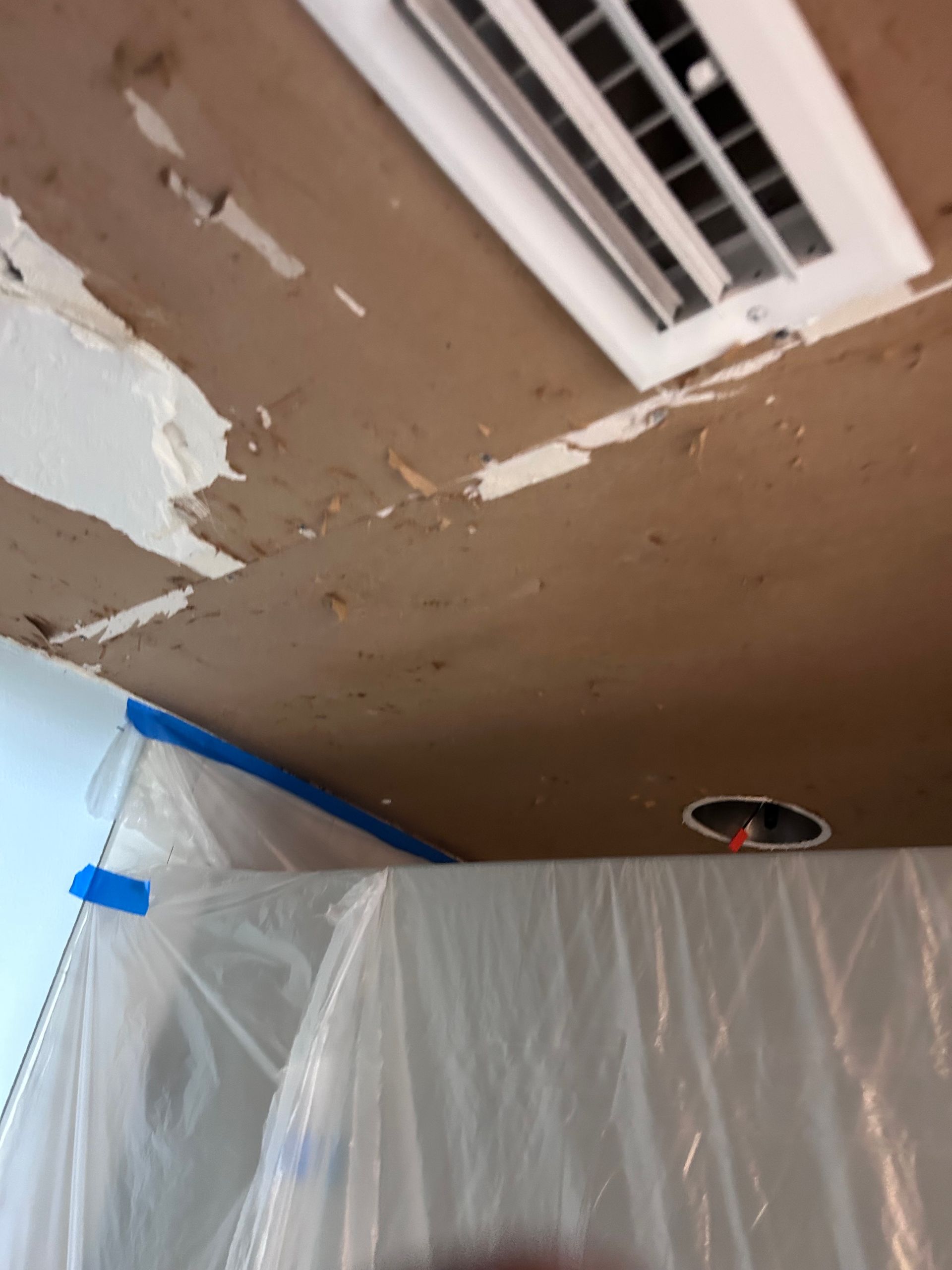 Ceiling with peeling paint, near a vent, covered in plastic, taped to blue wall.