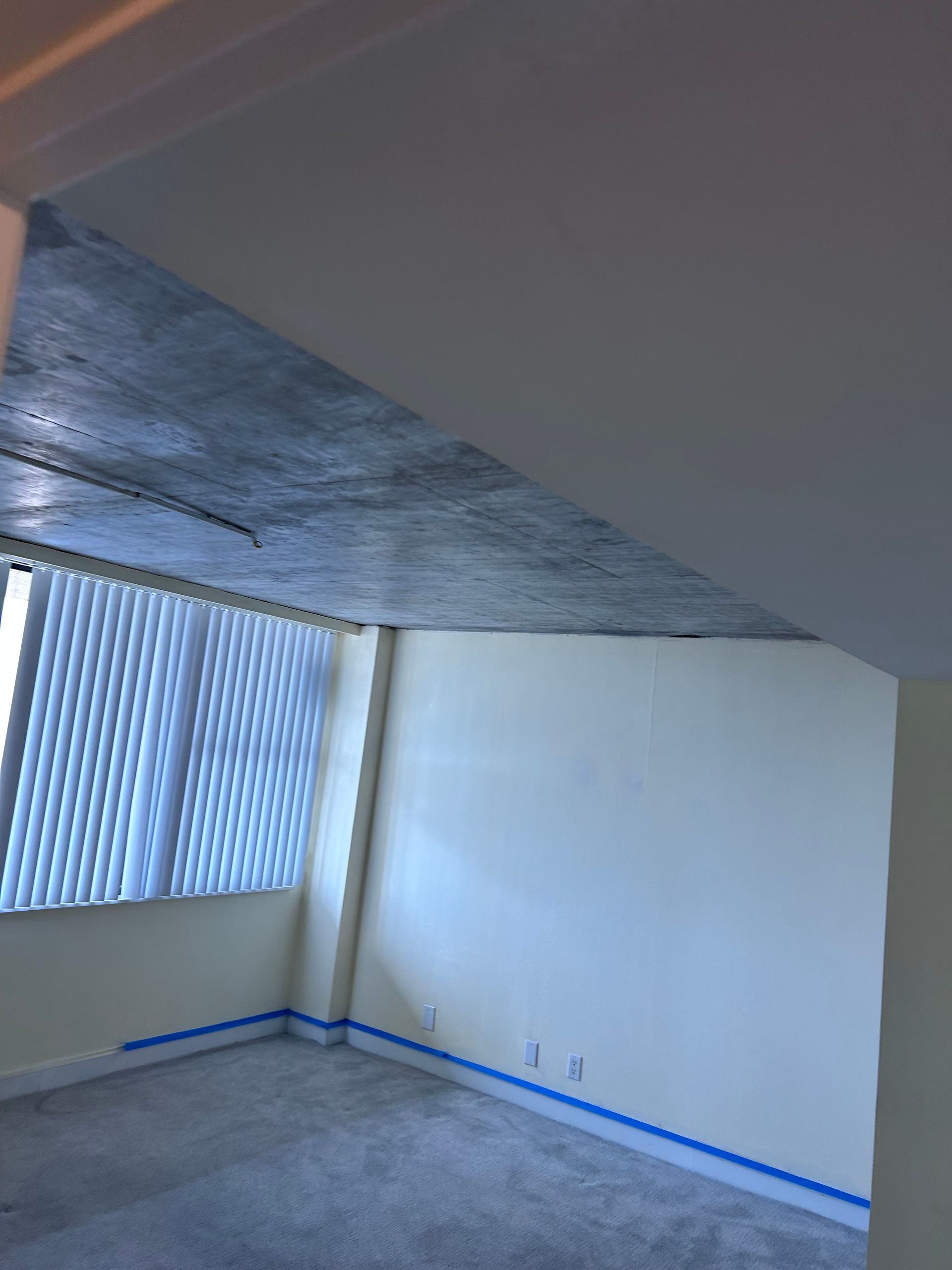 Empty room with concrete ceiling, large window, beige walls, and blue tape along the floor.