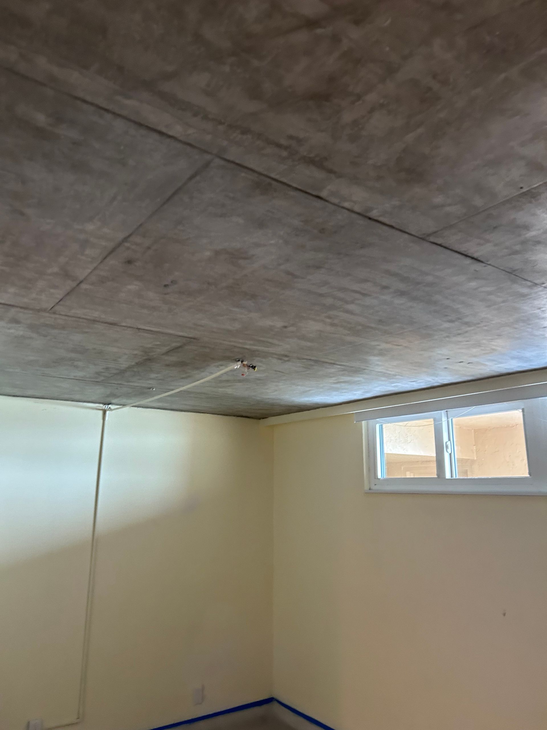 Basement corner with beige walls, concrete ceiling, and a small window with a white frame and white blinds.