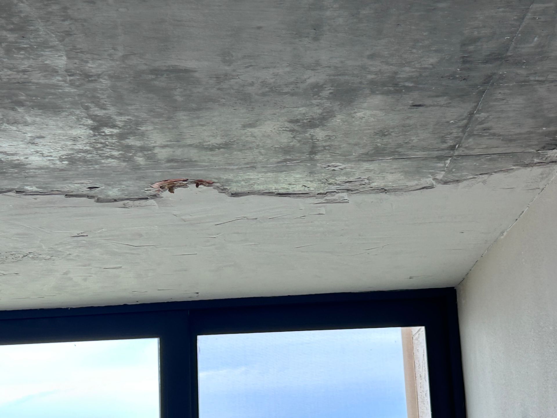 Cracked and damaged concrete ceiling above a dark window with a blue sky visible.