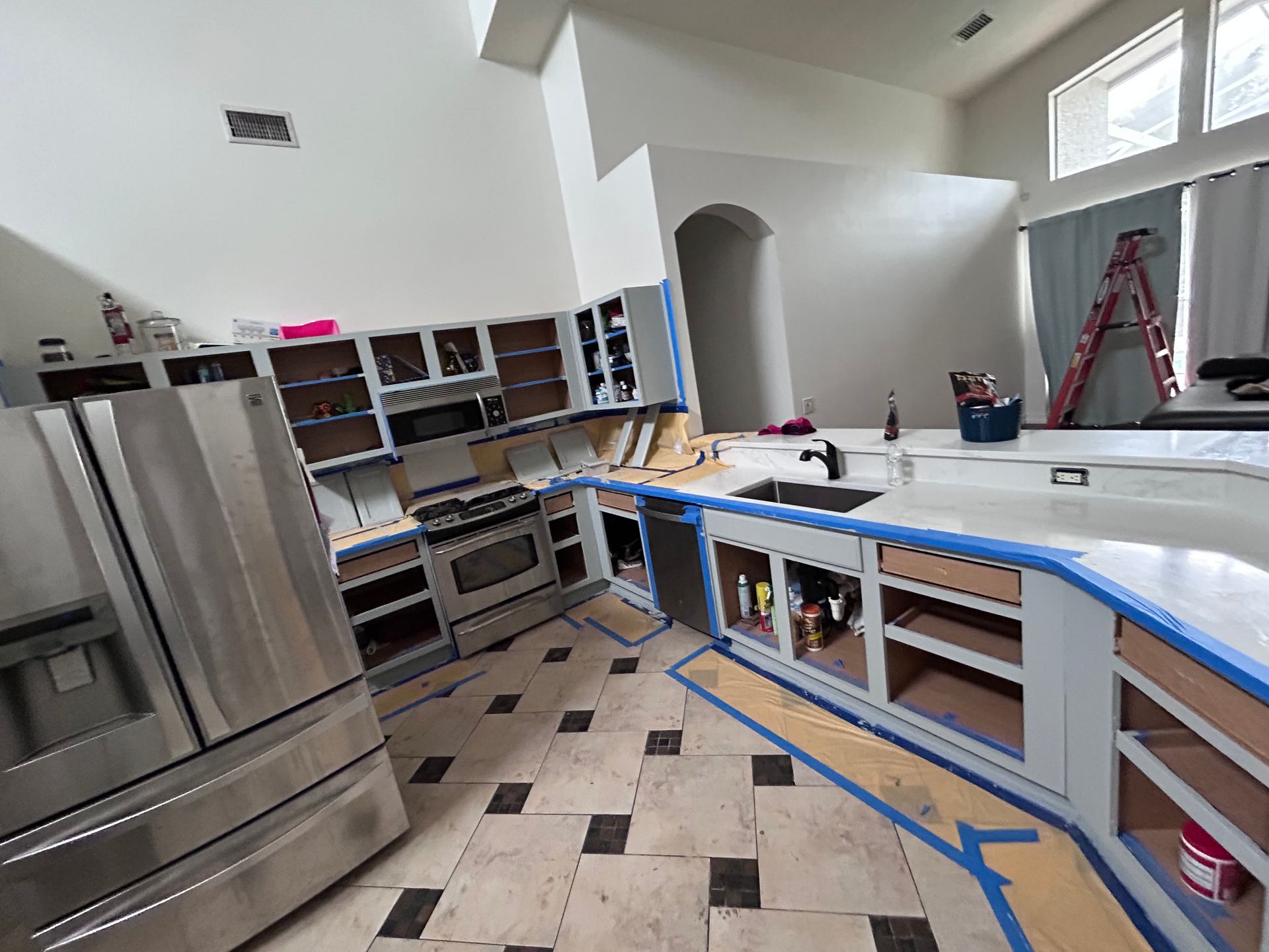 Kitchen under renovation with cabinets taped off for painting. Stainless steel appliances, tile floor.