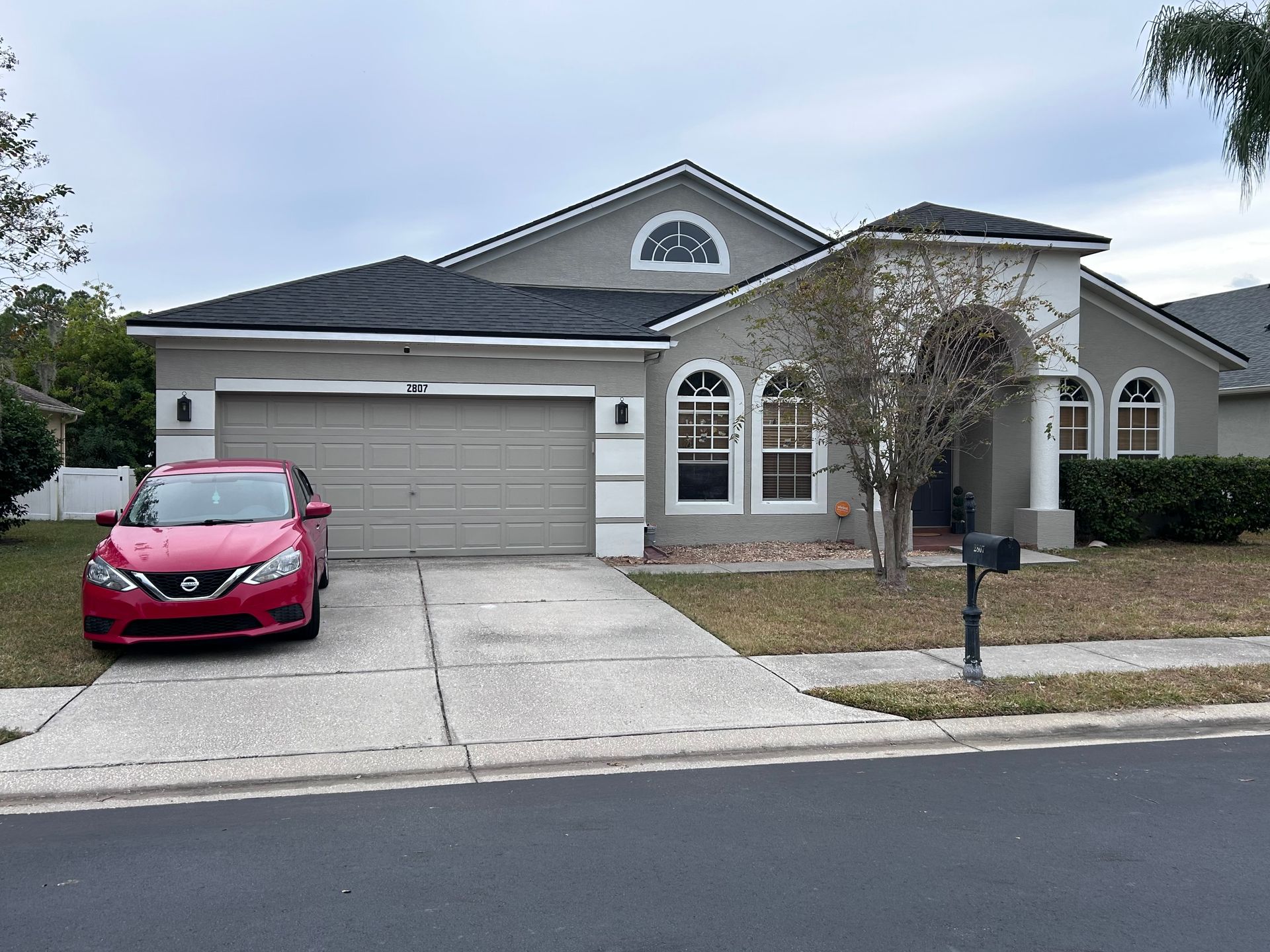 Red car parked in driveway of a two-story beige house with a dark roof and a small front yard.