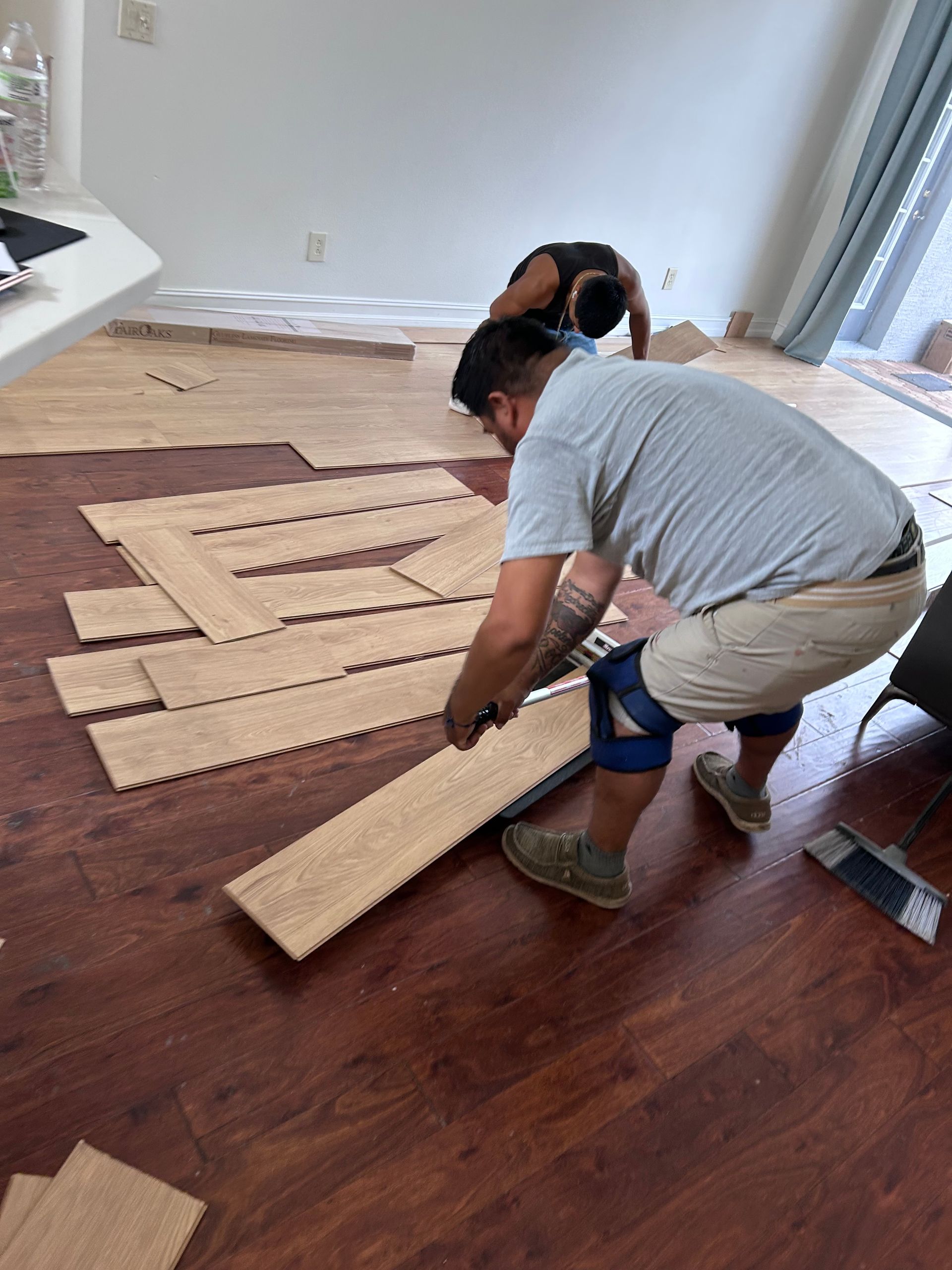 Two people installing wood flooring in a room with white walls, one is cutting planks, the other is laying them down.