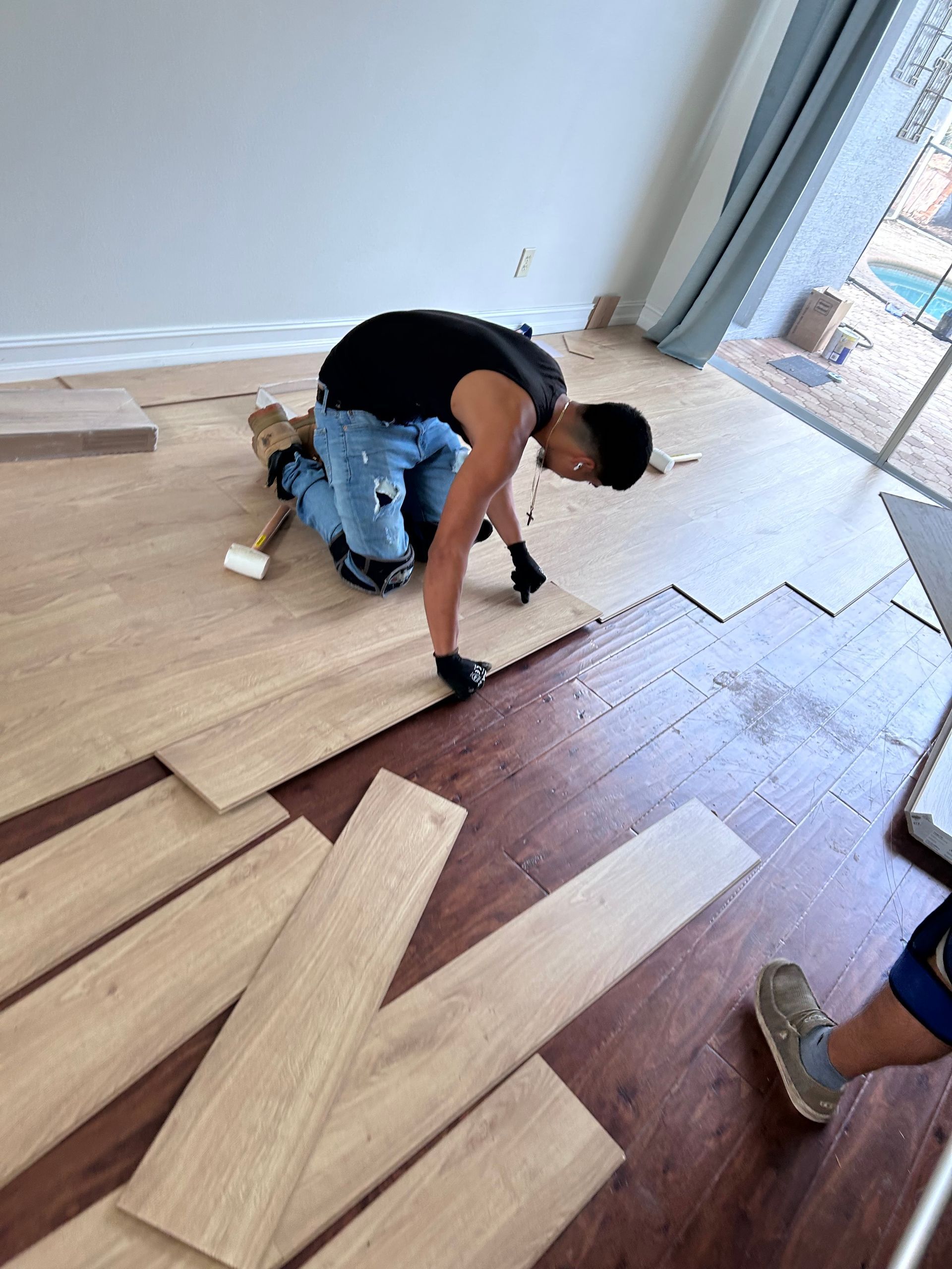 A person installs flooring, kneeling, placing a wood plank. Boards lay nearby on a floor with window and wall in the background.