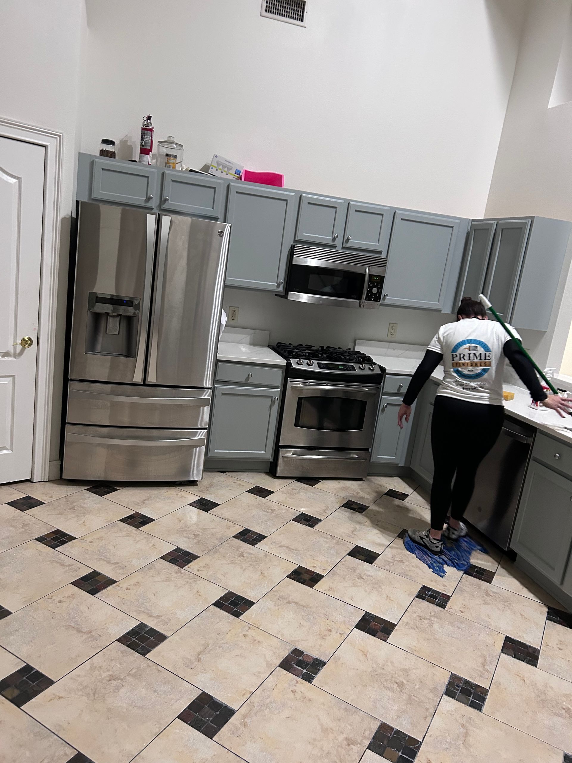 A person in a kitchen with gray cabinets, stainless steel appliances, and tiled floor.