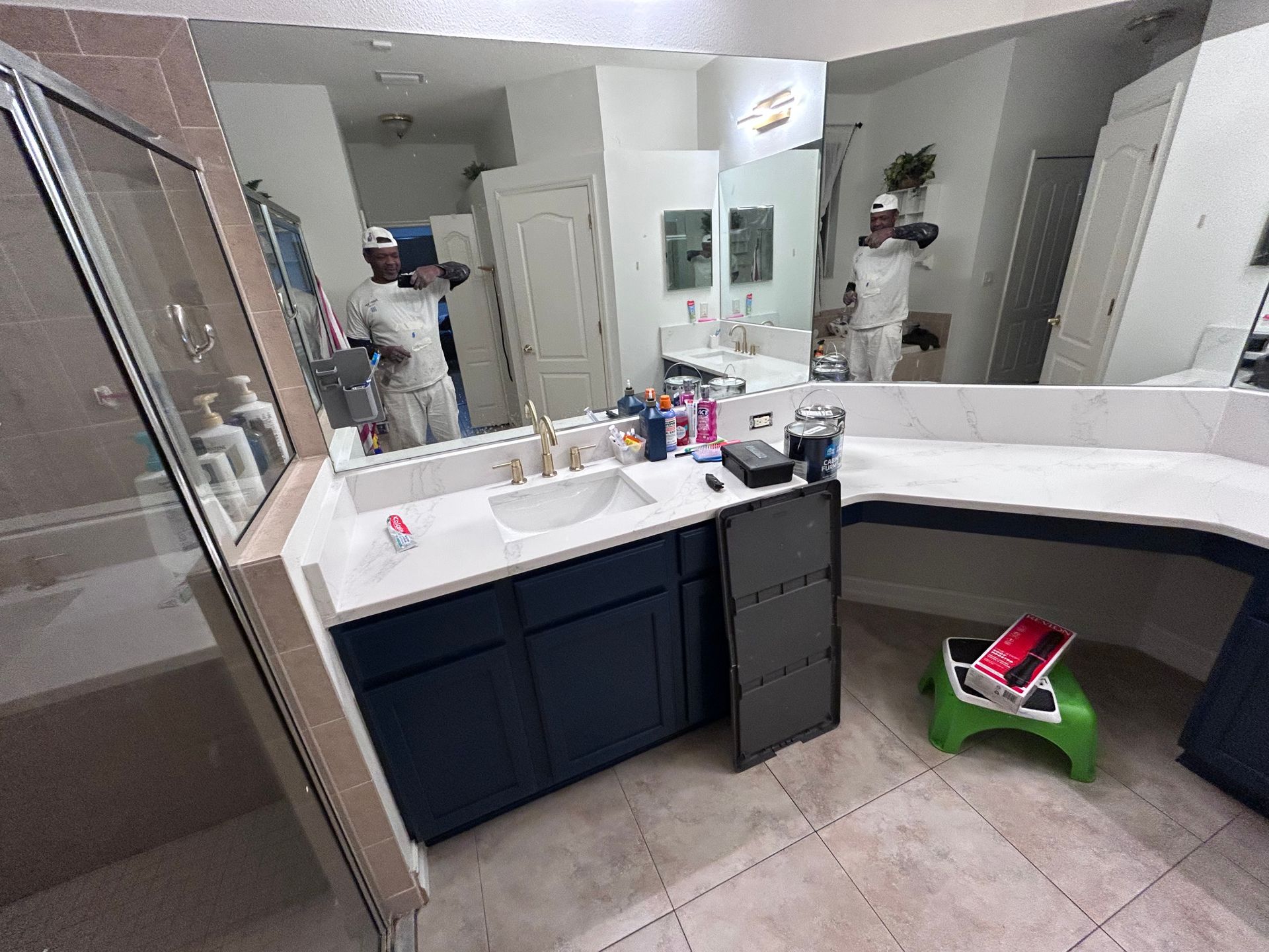 A person in a white outfit stands in a bathroom with a large mirror and blue cabinets.