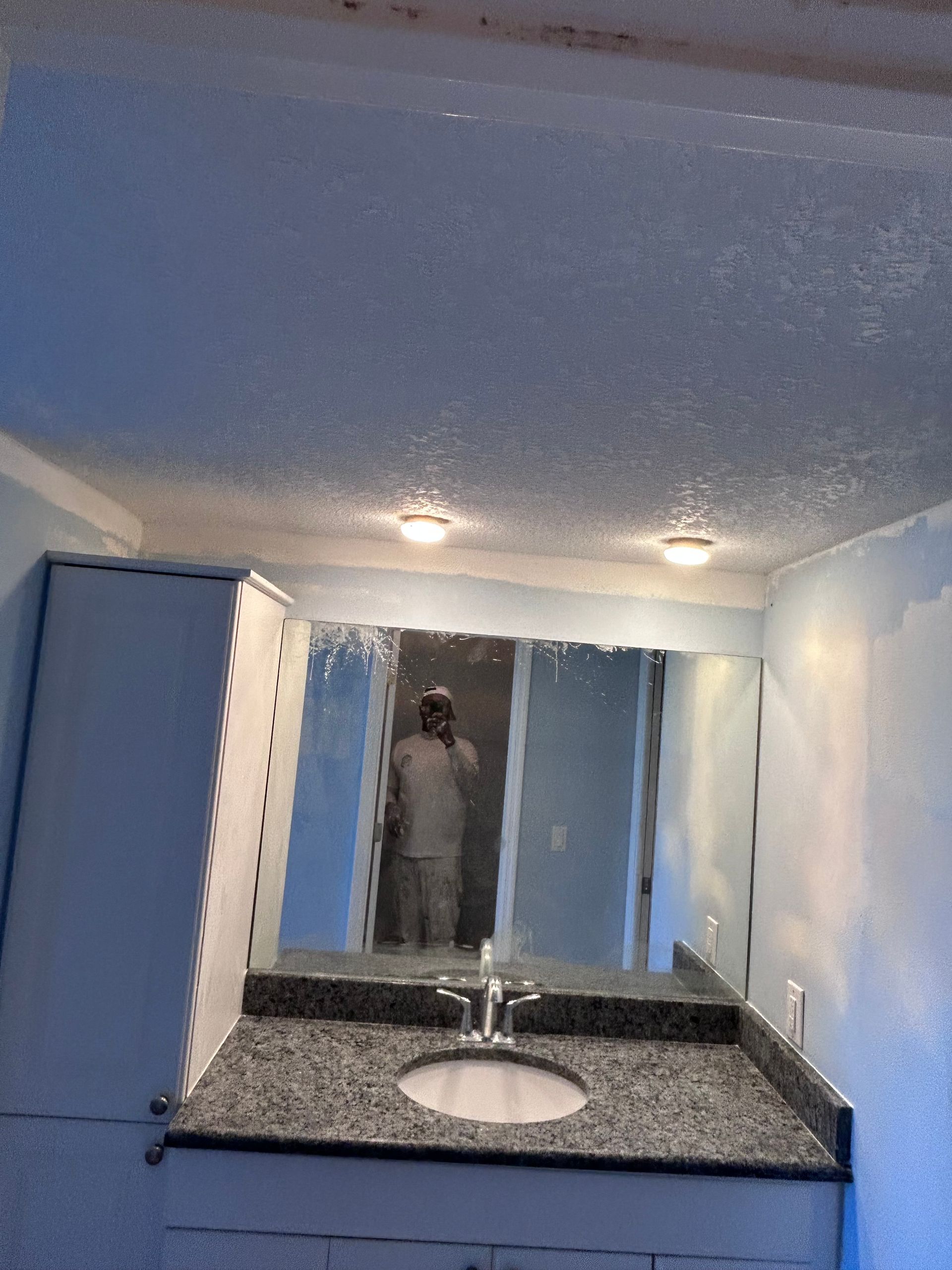Bathroom with a person reflected in the mirror, gray granite countertop, white sink, cabinets, and blue walls.
