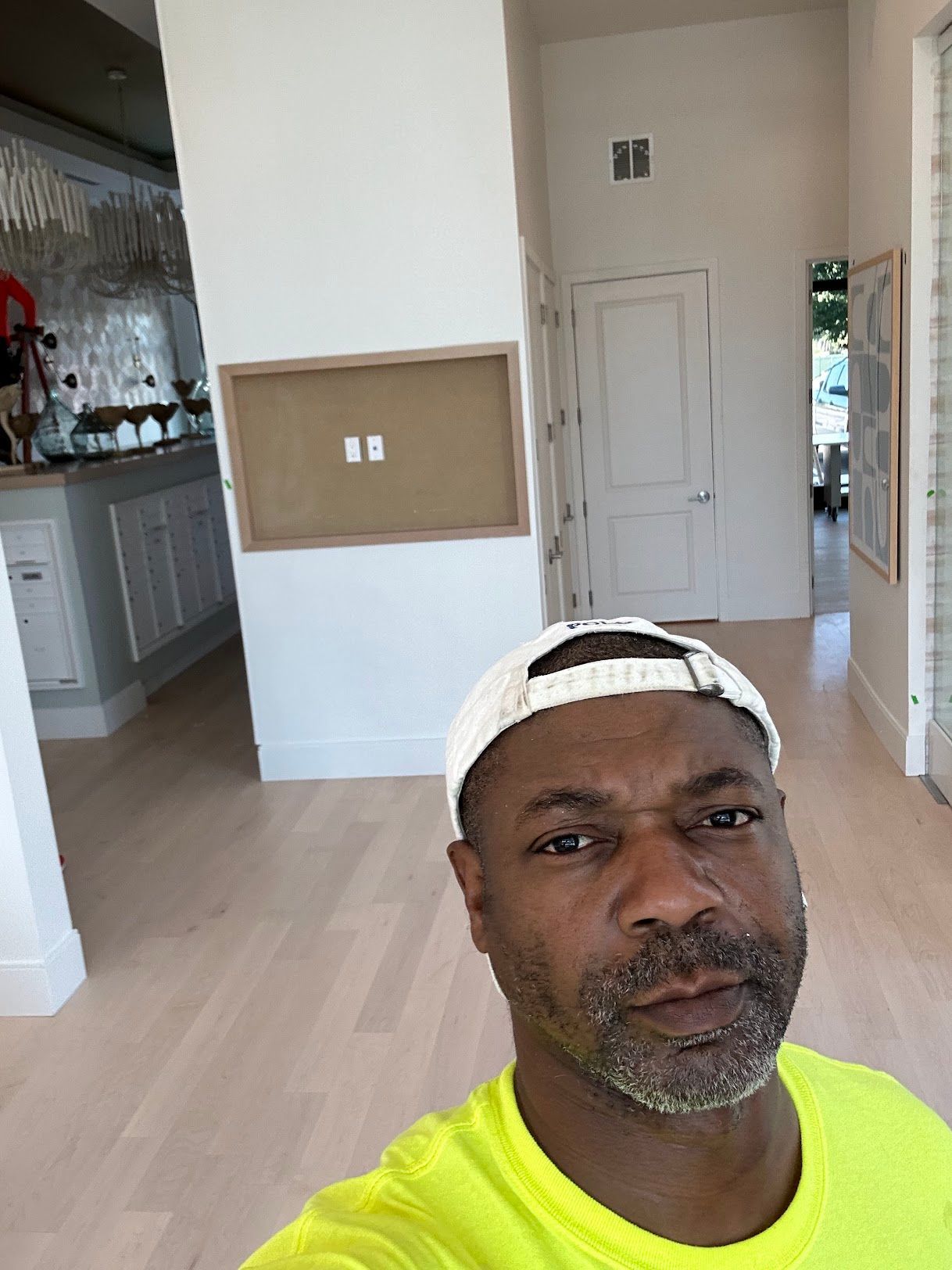 A man wearing a hat and a neon yellow shirt is taking a selfie in a hallway.
