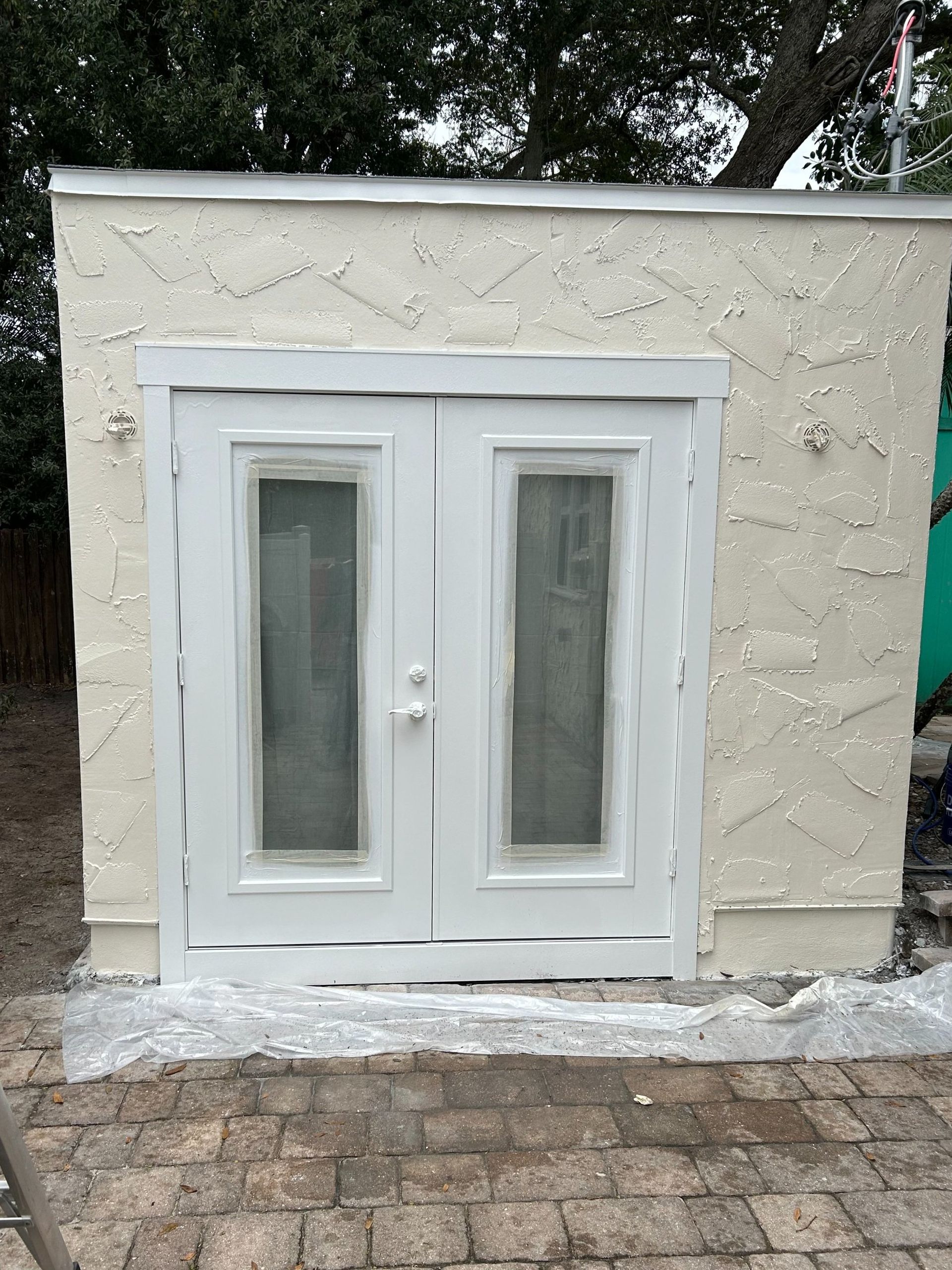 A white shed with two doors and two windows on a brick patio.