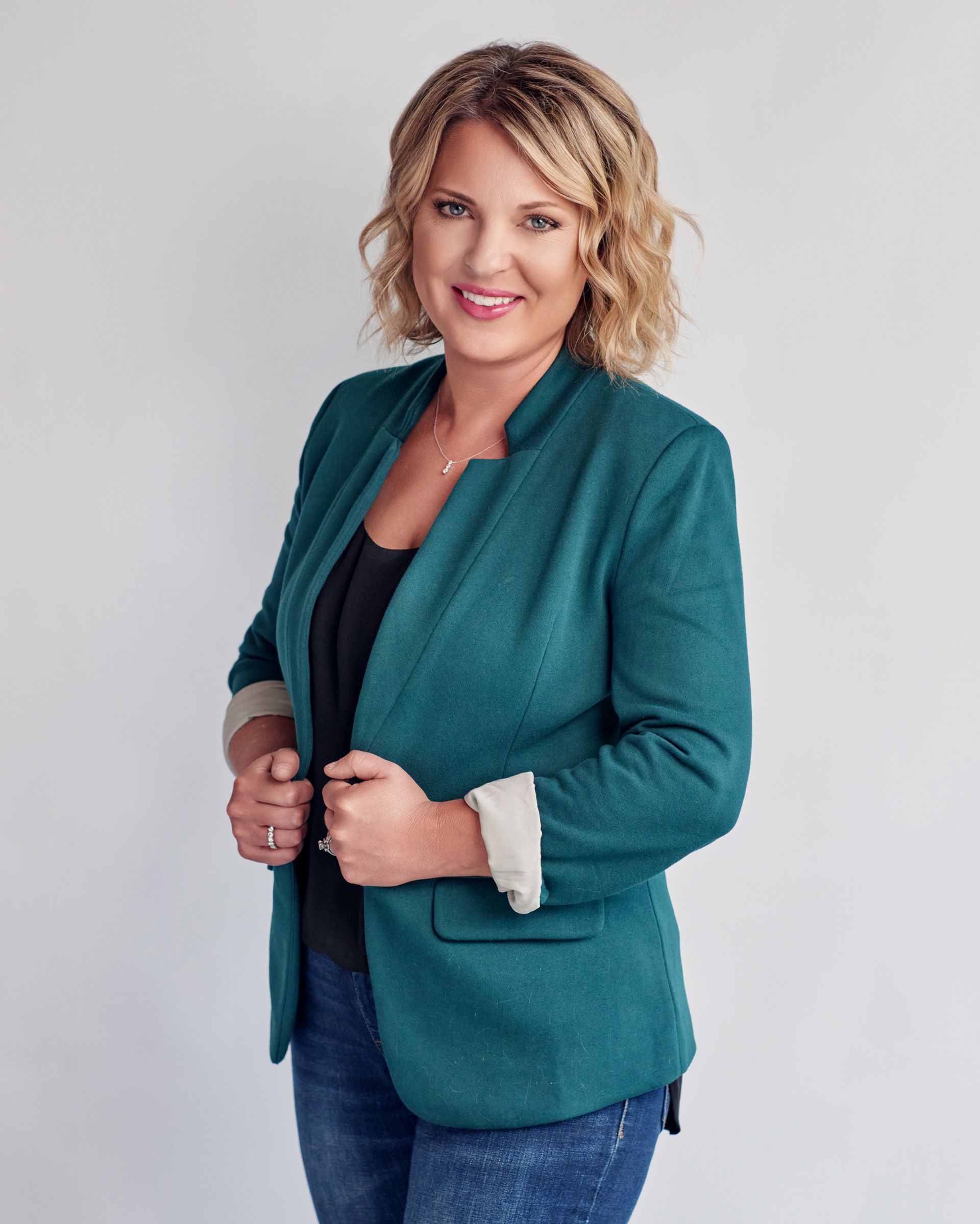 A woman wearing a green jacket and jeans is smiling for the camera.