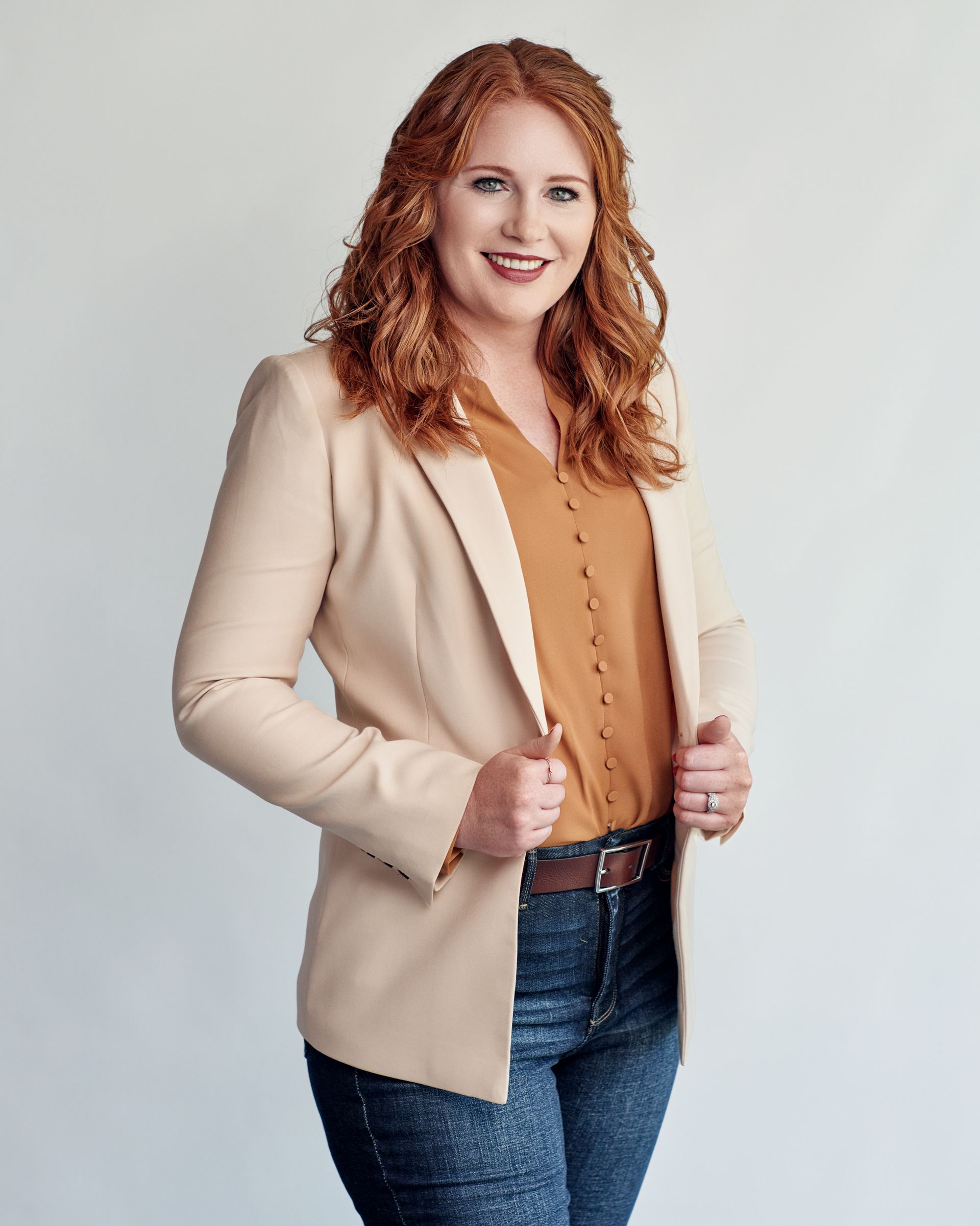 A woman with red hair is wearing a tan jacket and jeans.