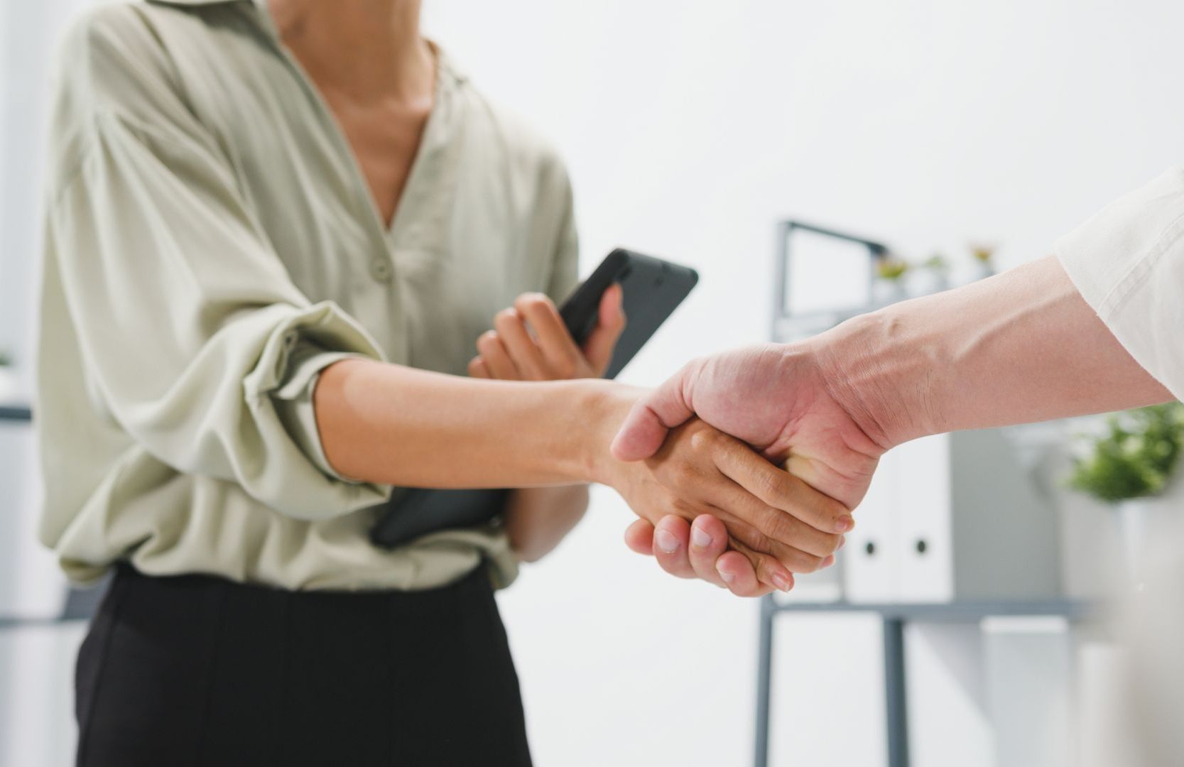 A woman is holding a cell phone while shaking hands with a man.